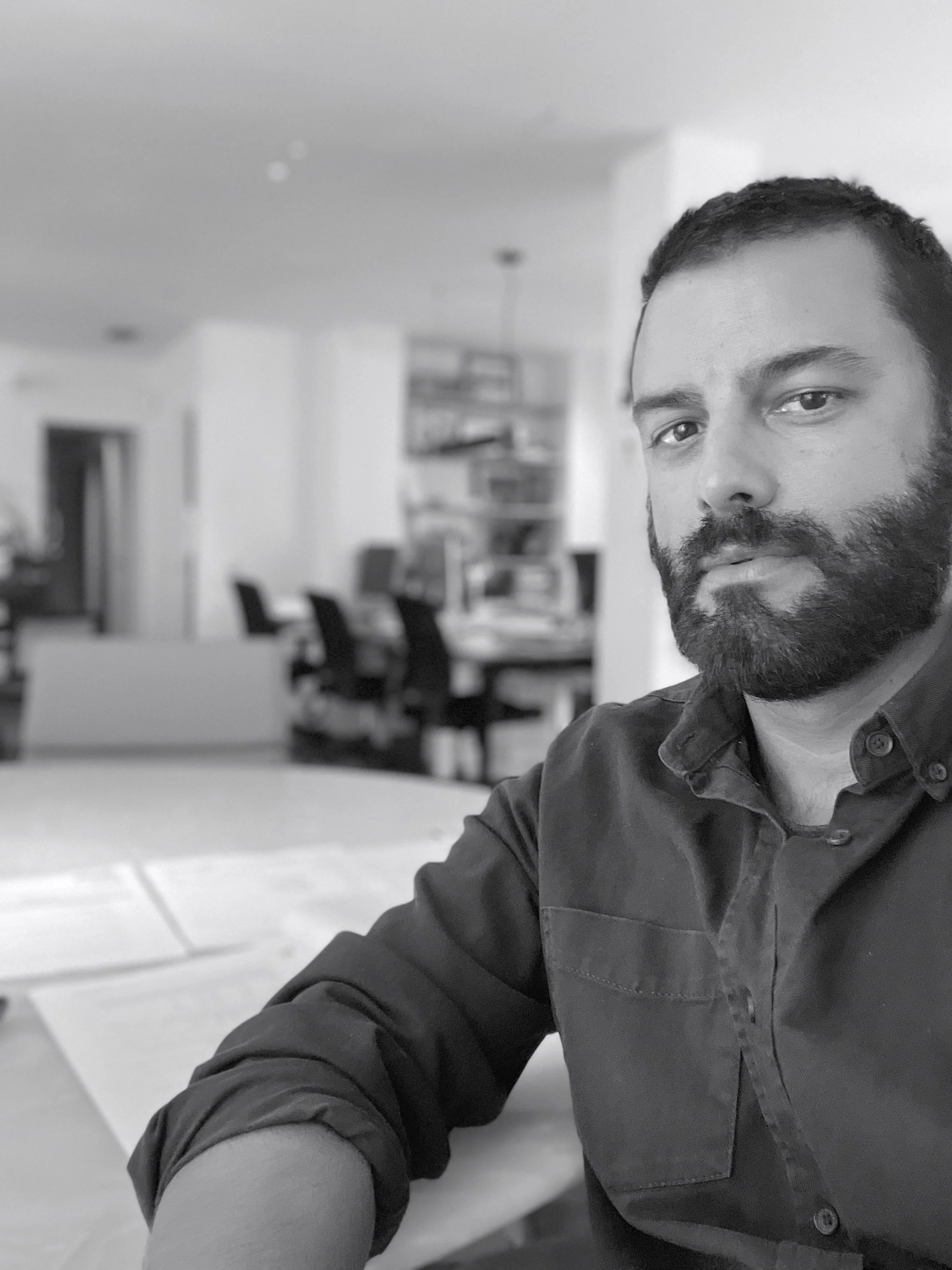 A man with a beard sitting at a table in a home or office setting, with a dining area and chairs in the background.