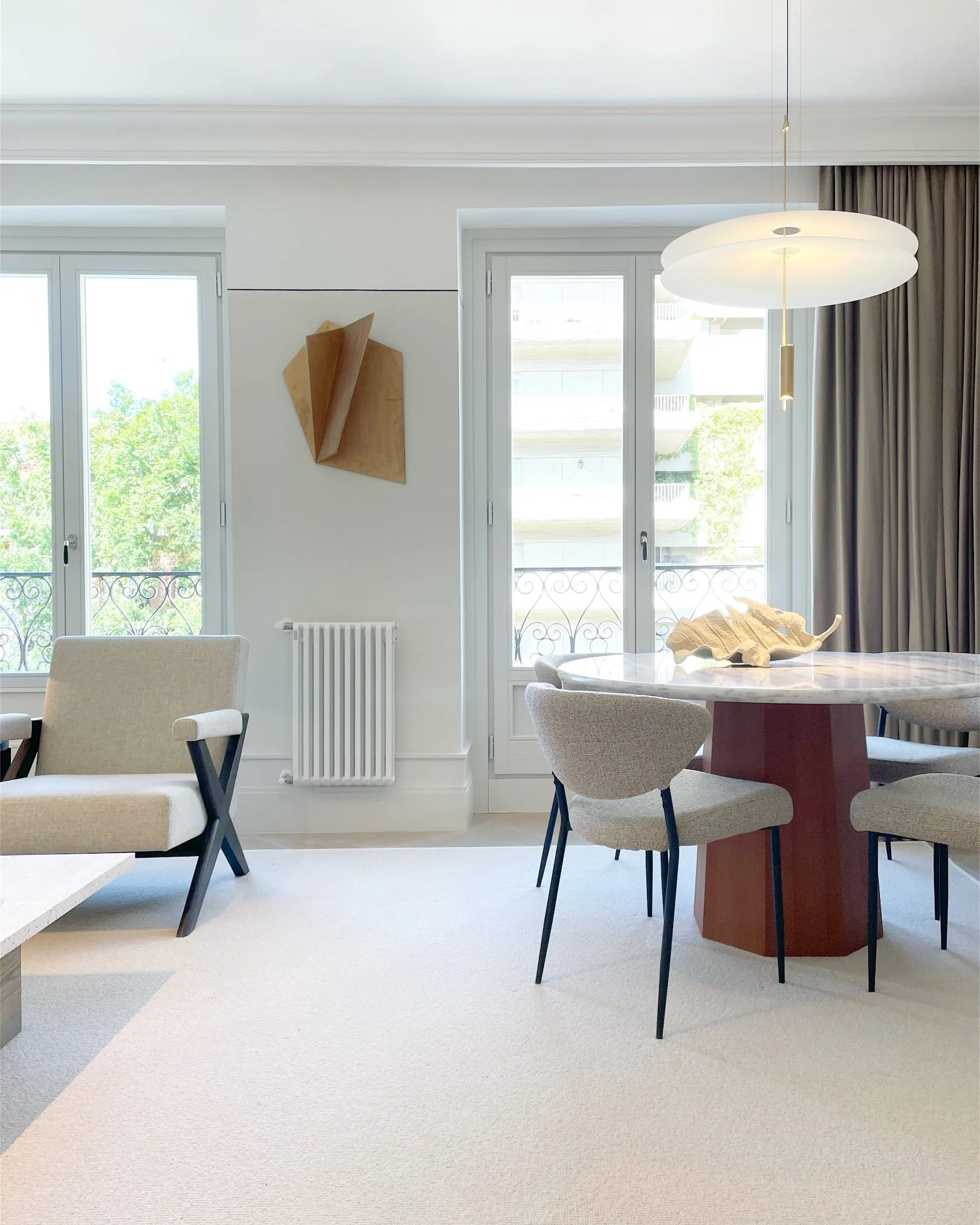 Modern living room with large windows, beige armchair, round marble dining table with beige chairs, abstract wooden wall art, ceiling light, and a beige carpet.