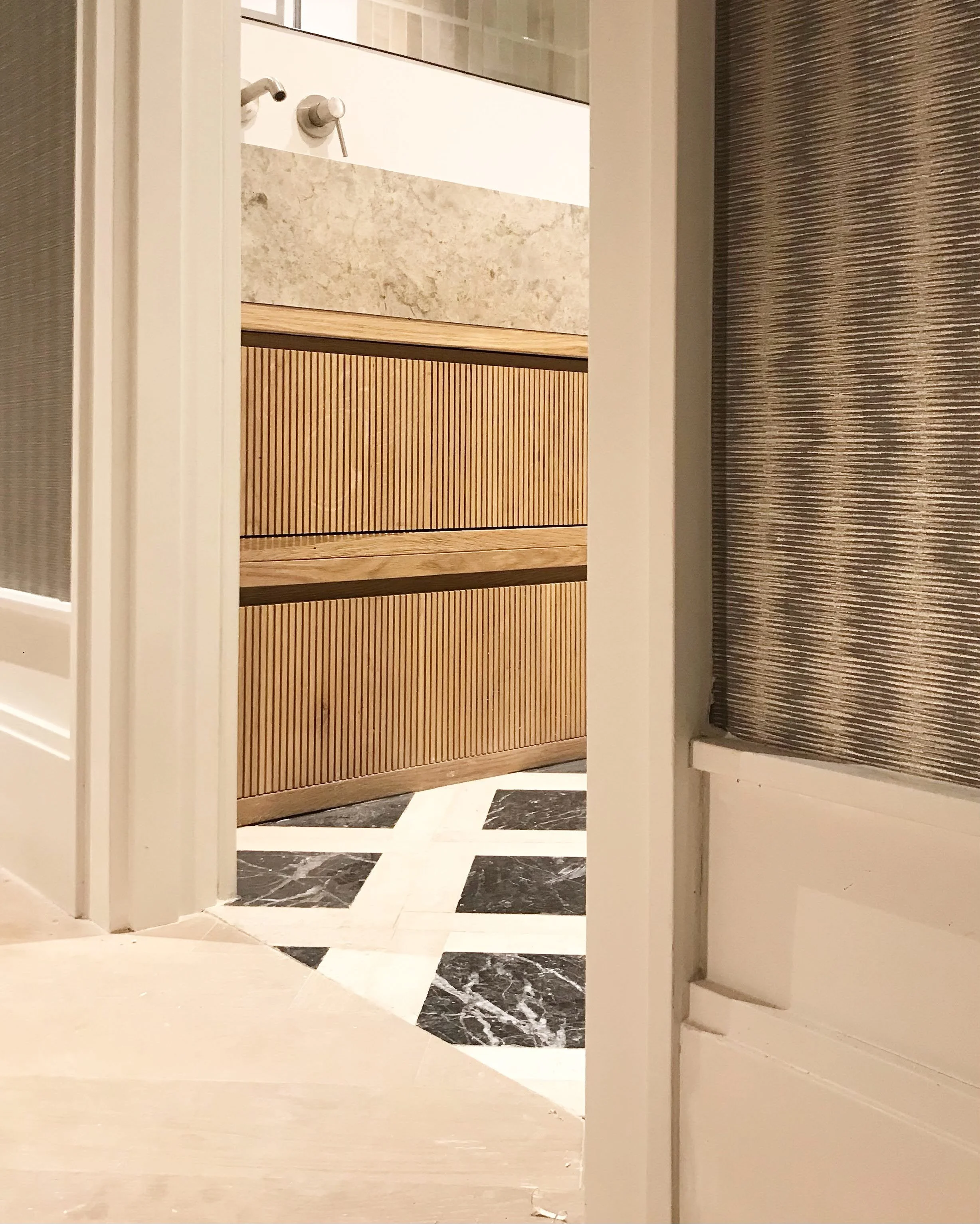 A view into a bathroom through an open door, showing a wooden cabinet with slatted drawers, a marble countertop, and a wall mirror. The floor has a black and white geometric pattern.