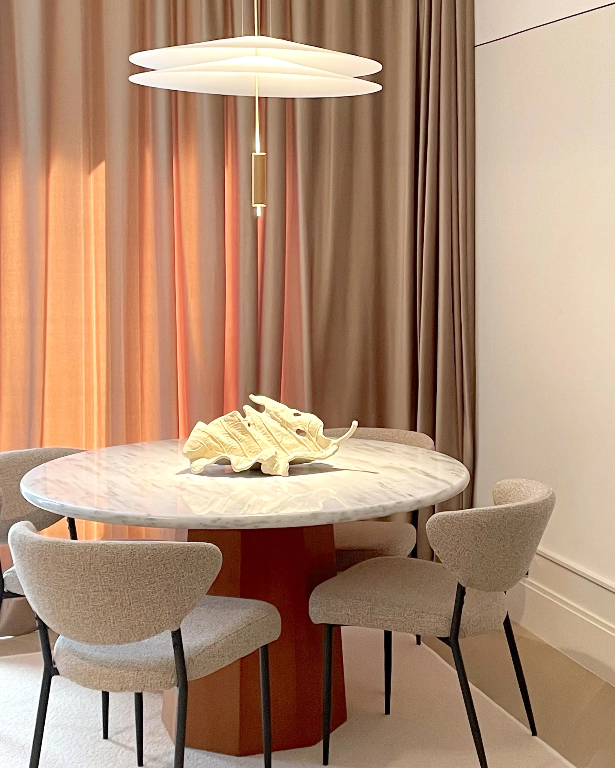 A round marble dining table with a decorative shell centerpiece, surrounded by four beige upholstered chairs, with a modern white pendant light hanging above and beige curtains in the background.