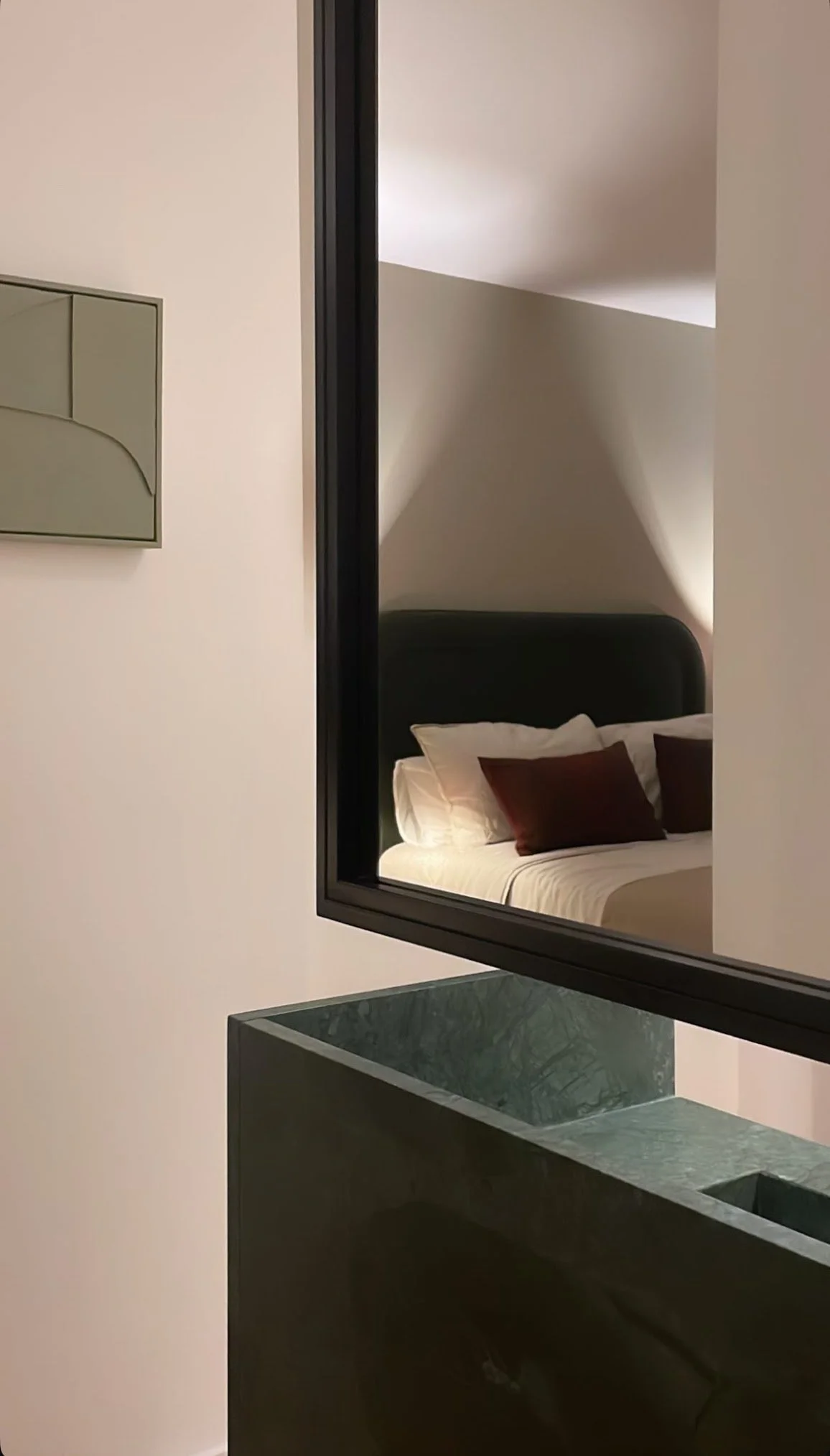 A modern hotel room with a bed reflected in a mirror. The bed has white linens and brown pillows. The room has a sloped ceiling and minimal decor.