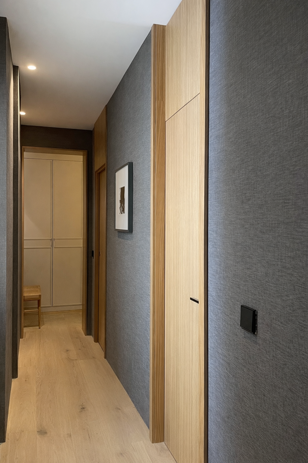 A narrow hallway with gray textured walls, wooden doors, and light wood flooring. There is a framed picture on the wall and a small wooden bench in a small alcove at the end.