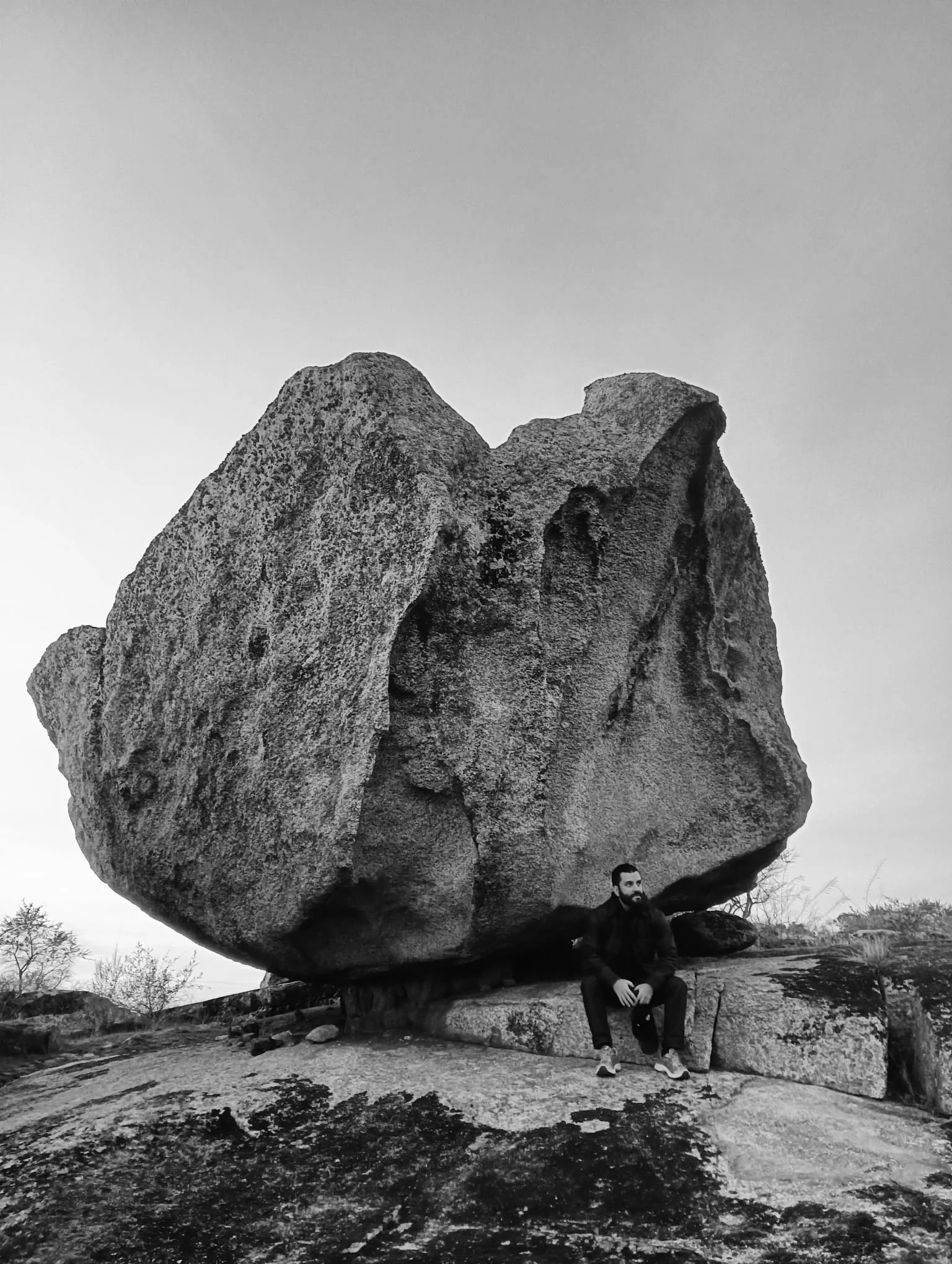 A man with a beard and dark clothing sitting on rock underneath a large, irregularly shaped boulder.