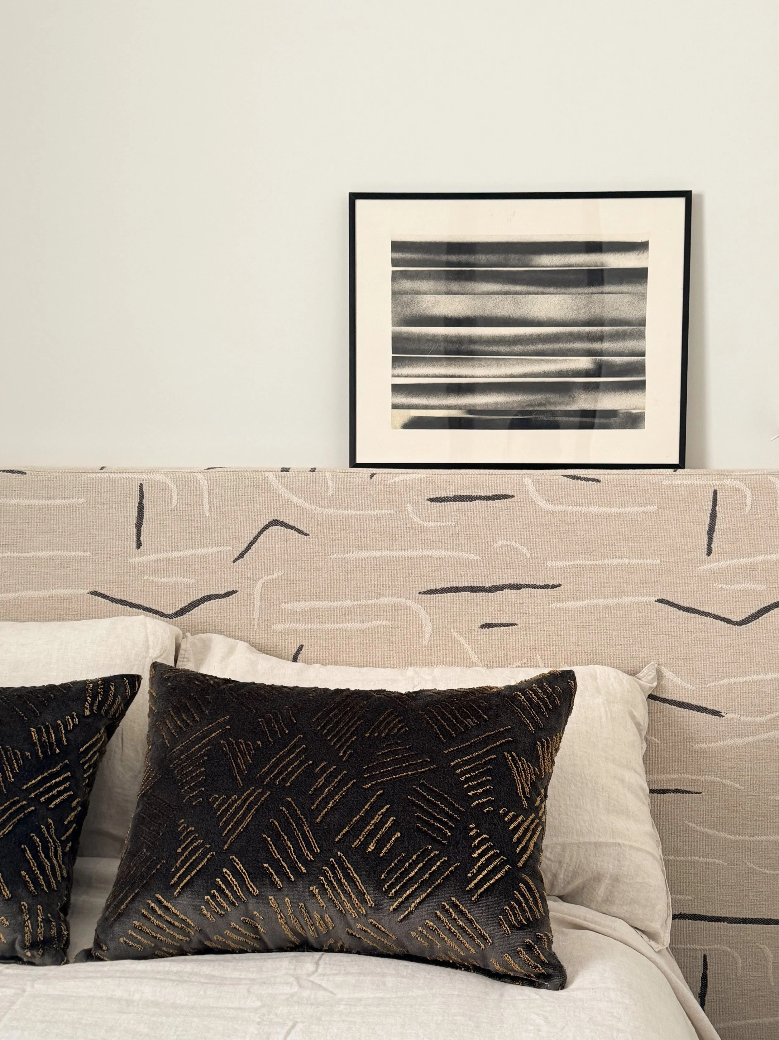 A bedroom scene with a beige upholstered headboard with black abstract lines, two black pillows with gold embroidery, a white pillow, and a framed black and white striped artwork on a plain white wall.