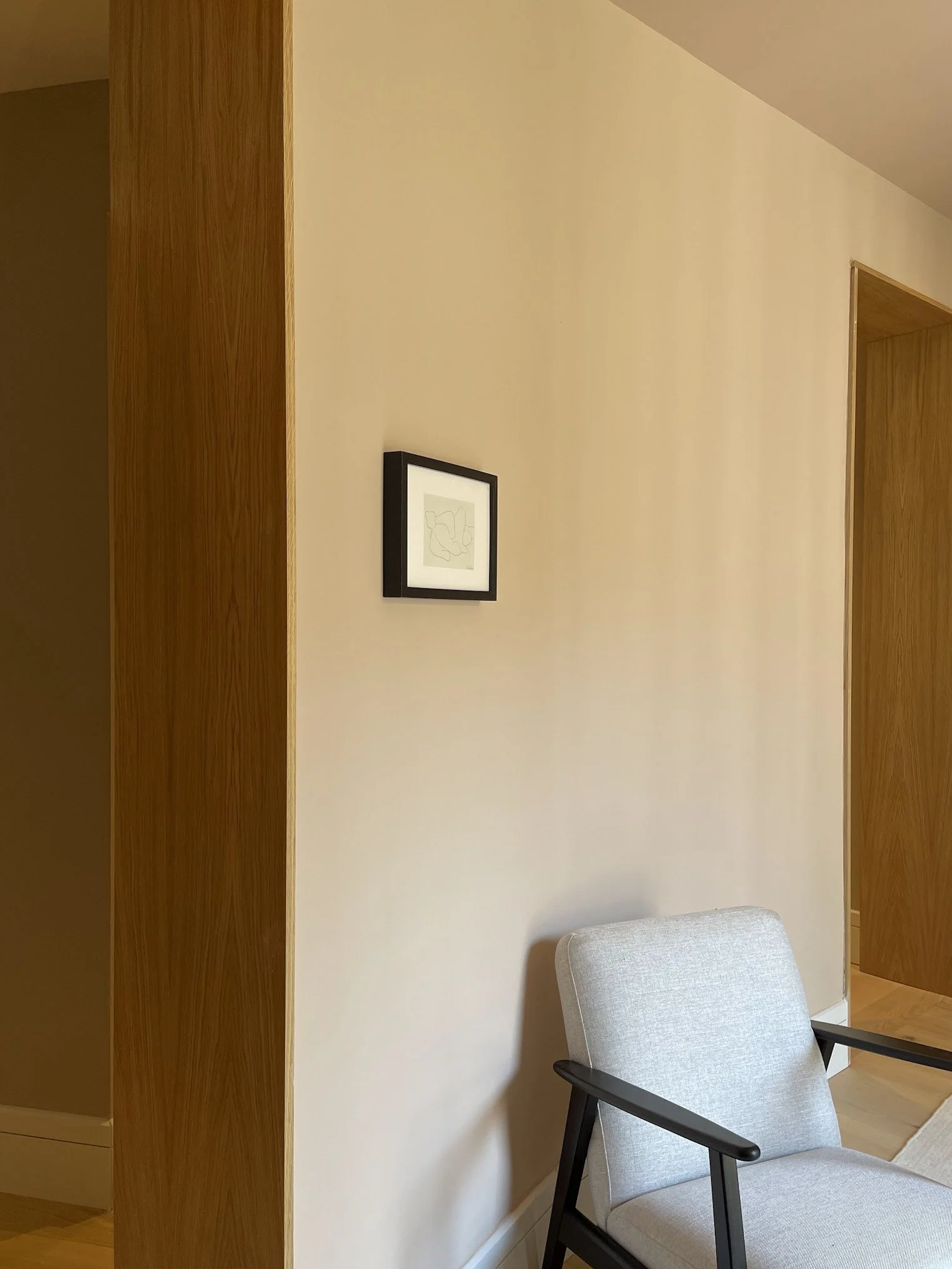 A corner of a room with a beige wall, a framed abstract line drawing, a gray upholstered armchair with black armrests, and wooden accents.