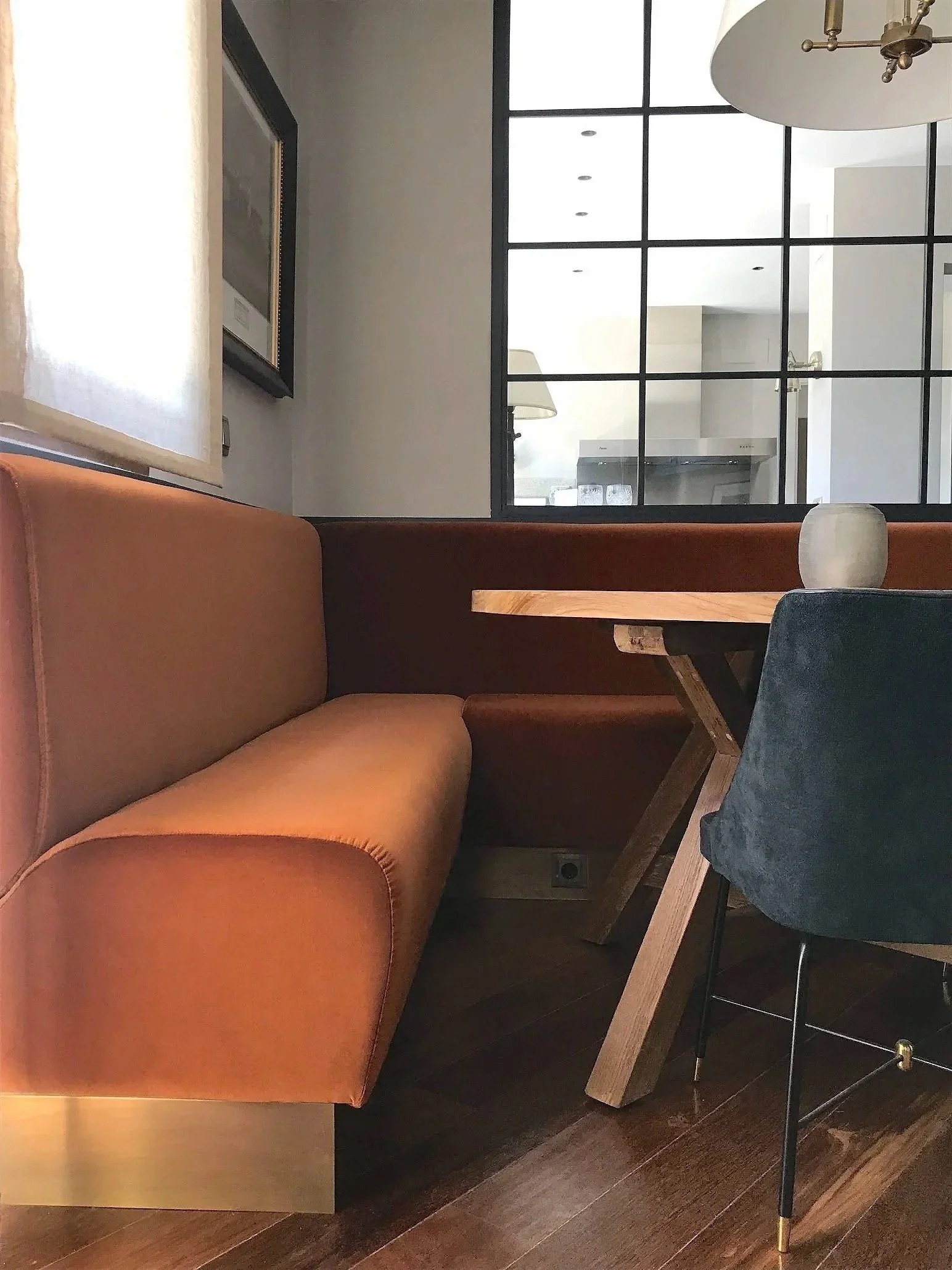 Interior of a restaurant with a brown leather booth, a wooden table, a blue chair, and a decorative vase, with a large window and glass partition walls in the background.