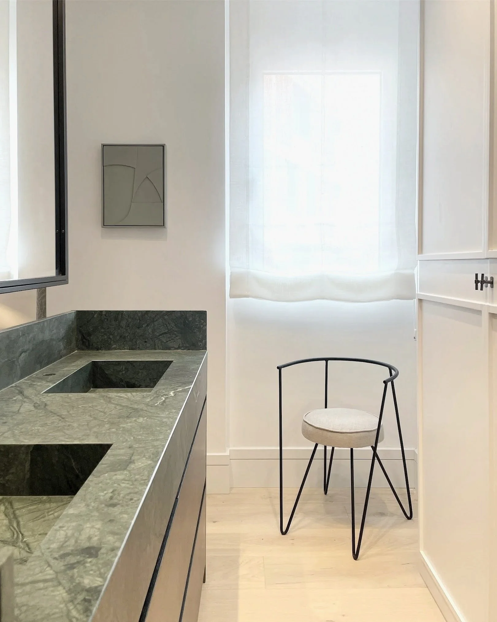 Modern kitchen with marble countertop and a white curtain covering a window, a decorative art piece on the wall, and a round stool with a metal frame.
