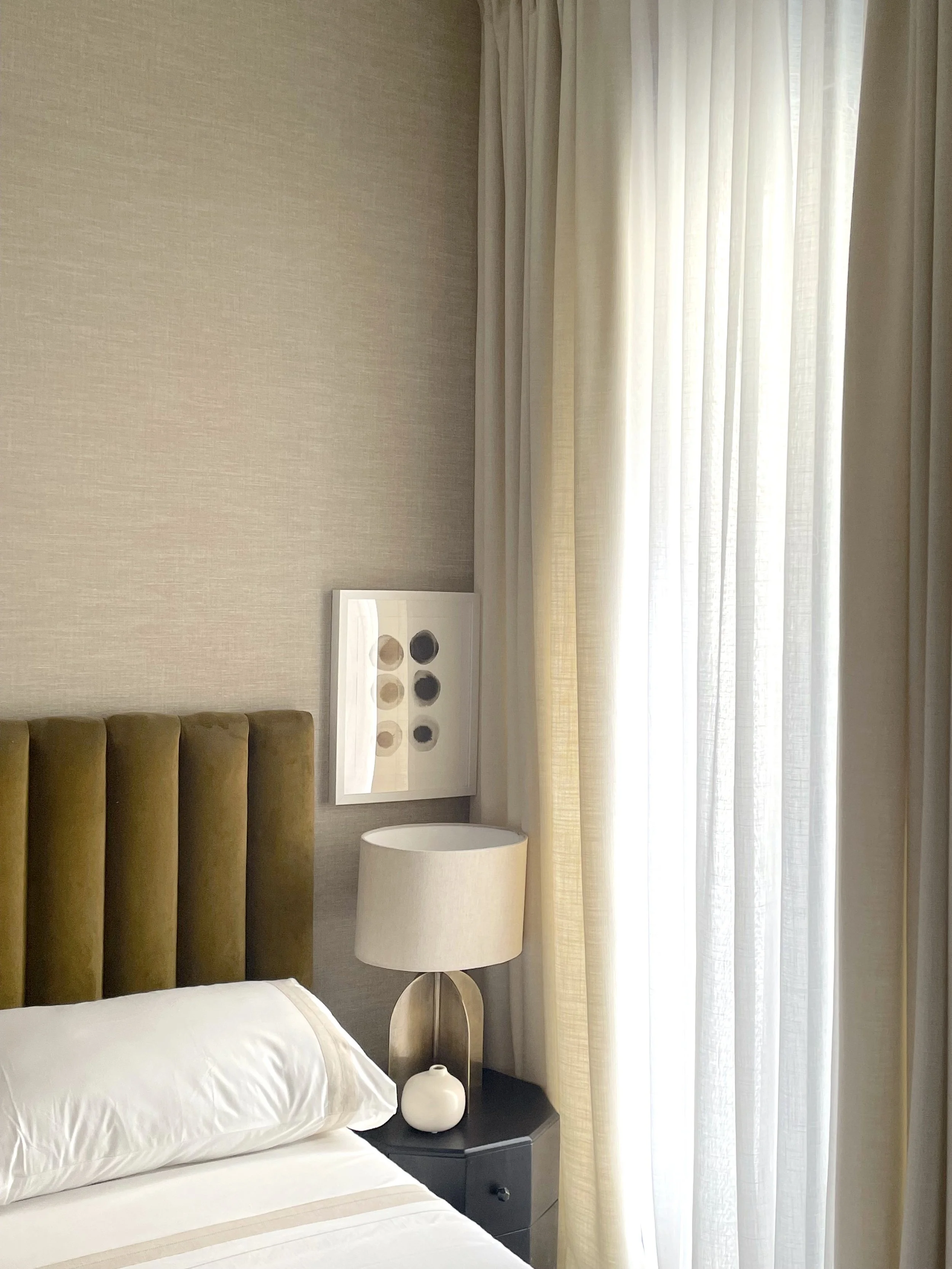 A bedroom with beige walls, a bed with white bedding and pillows, a brown upholstered headboard, a nightstand with a beige lamp and white vase, and a window with sheer white curtains.