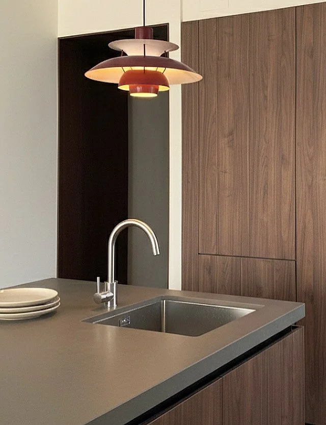 Modern kitchen with a gray countertop, stainless steel sink, and a chrome faucet. There is a stack of white plates, wooden cabinets, and a colorful layered pendant light hanging above.