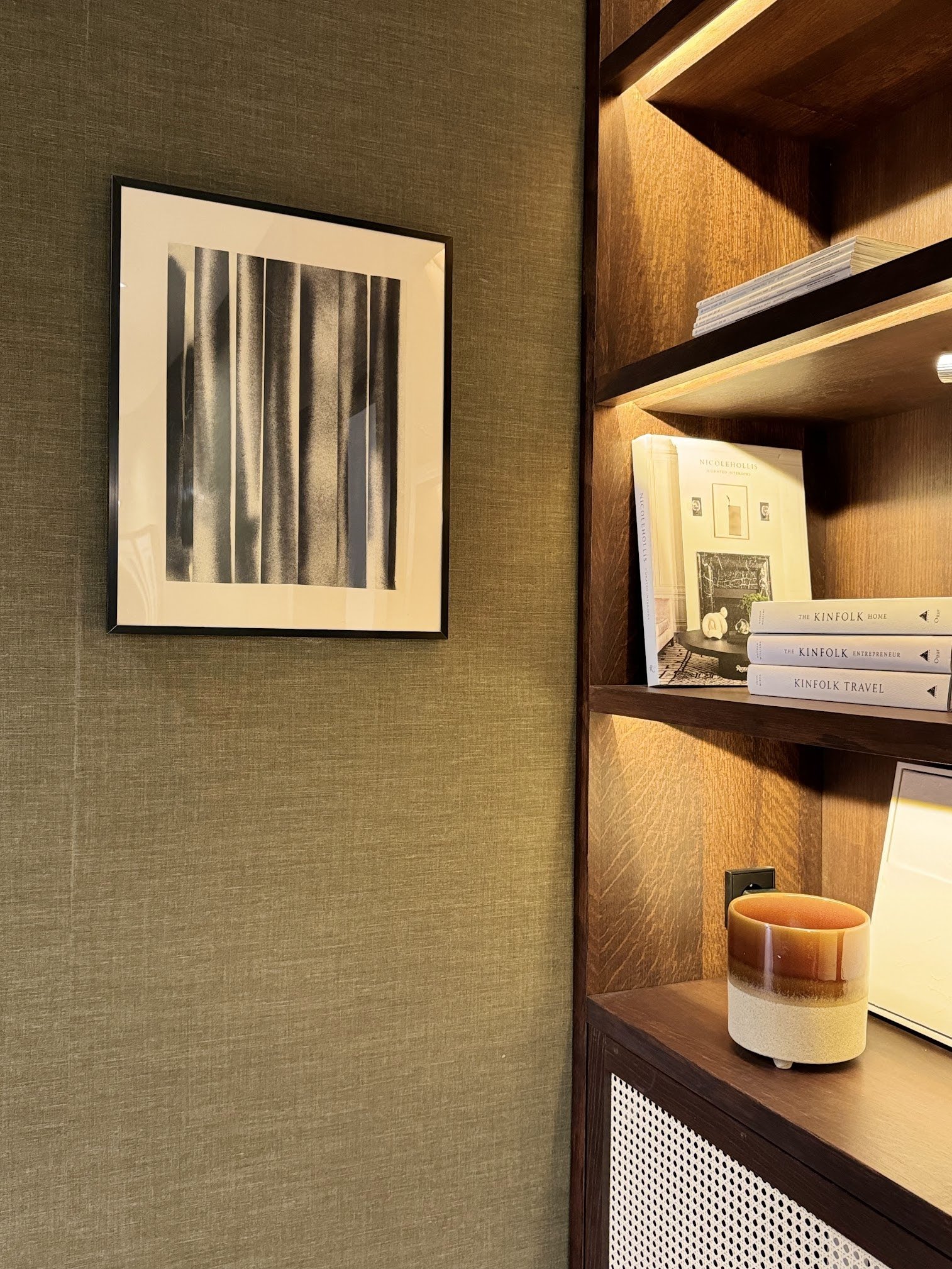 A framed black-and-white modern art print hanging on a textured beige wall next to a wooden bookshelf with books and decorative objects.
