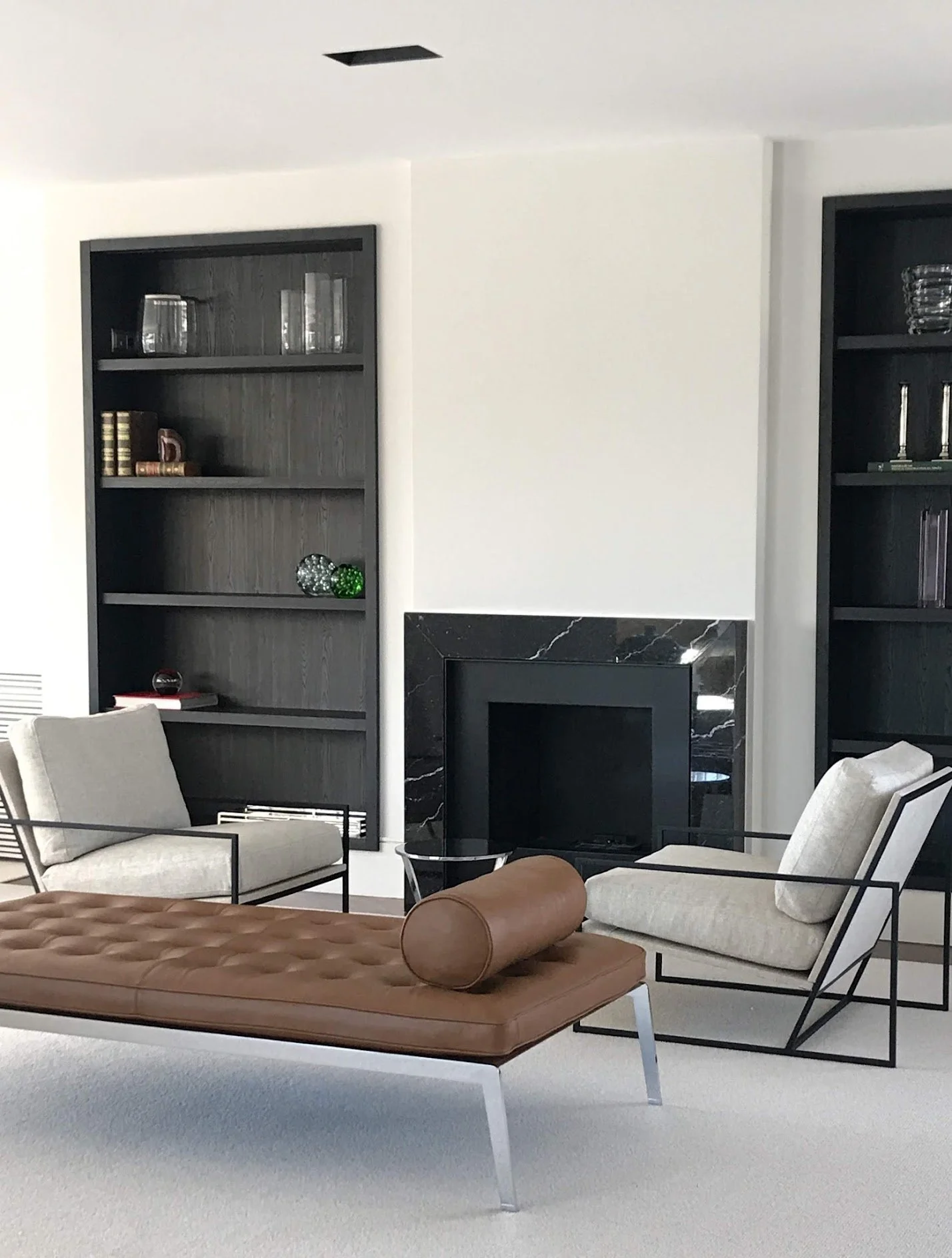 Modern living room with a fireplace, black bookshelves, two beige armchairs, and a brown leather bench with a cylindrical pillow.