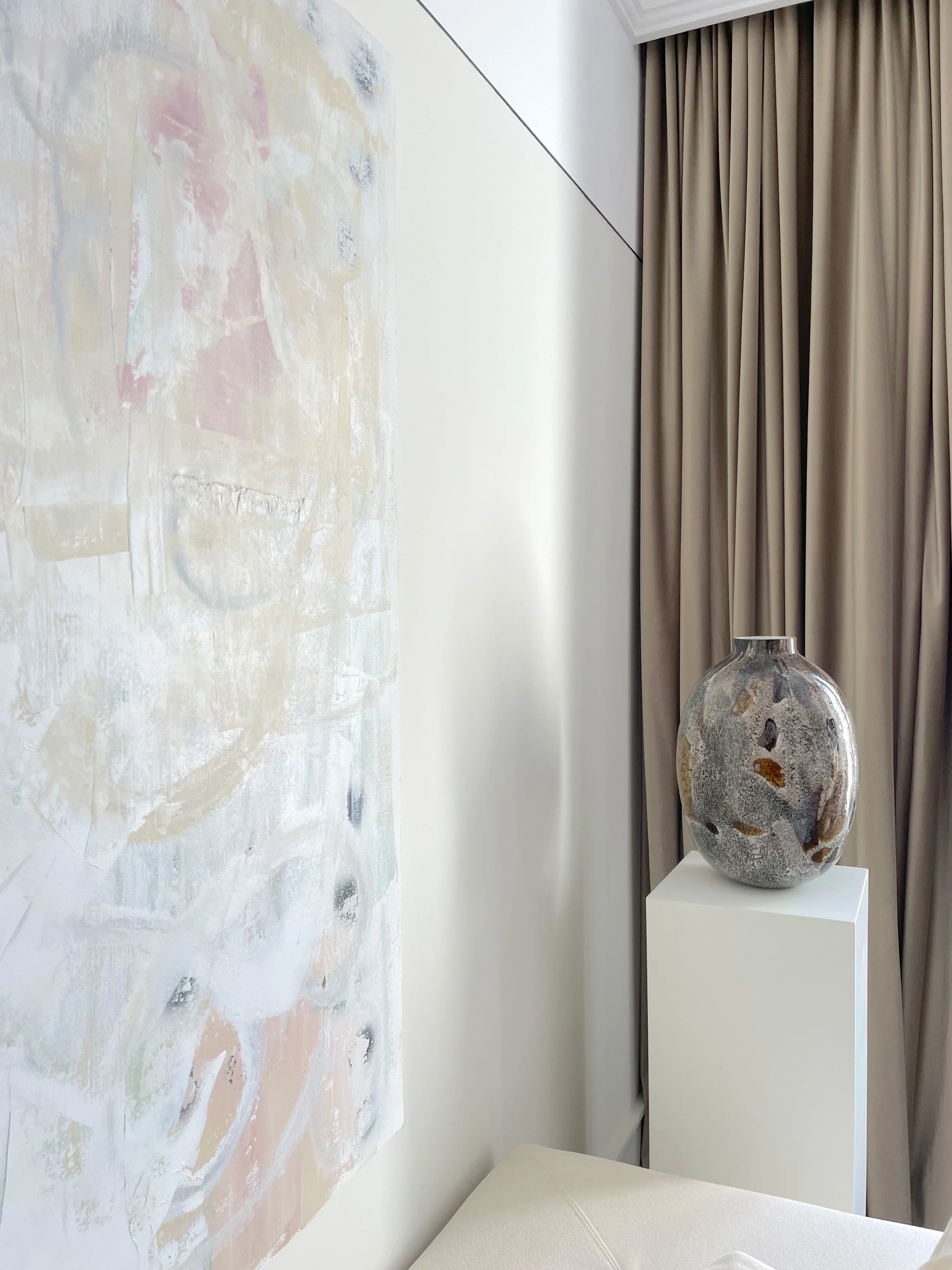 A decorative abstract painting on the wall and a large, rounded glass vase with an earthy, textured design placed on a white pedestal next to beige curtains in a modern interior.