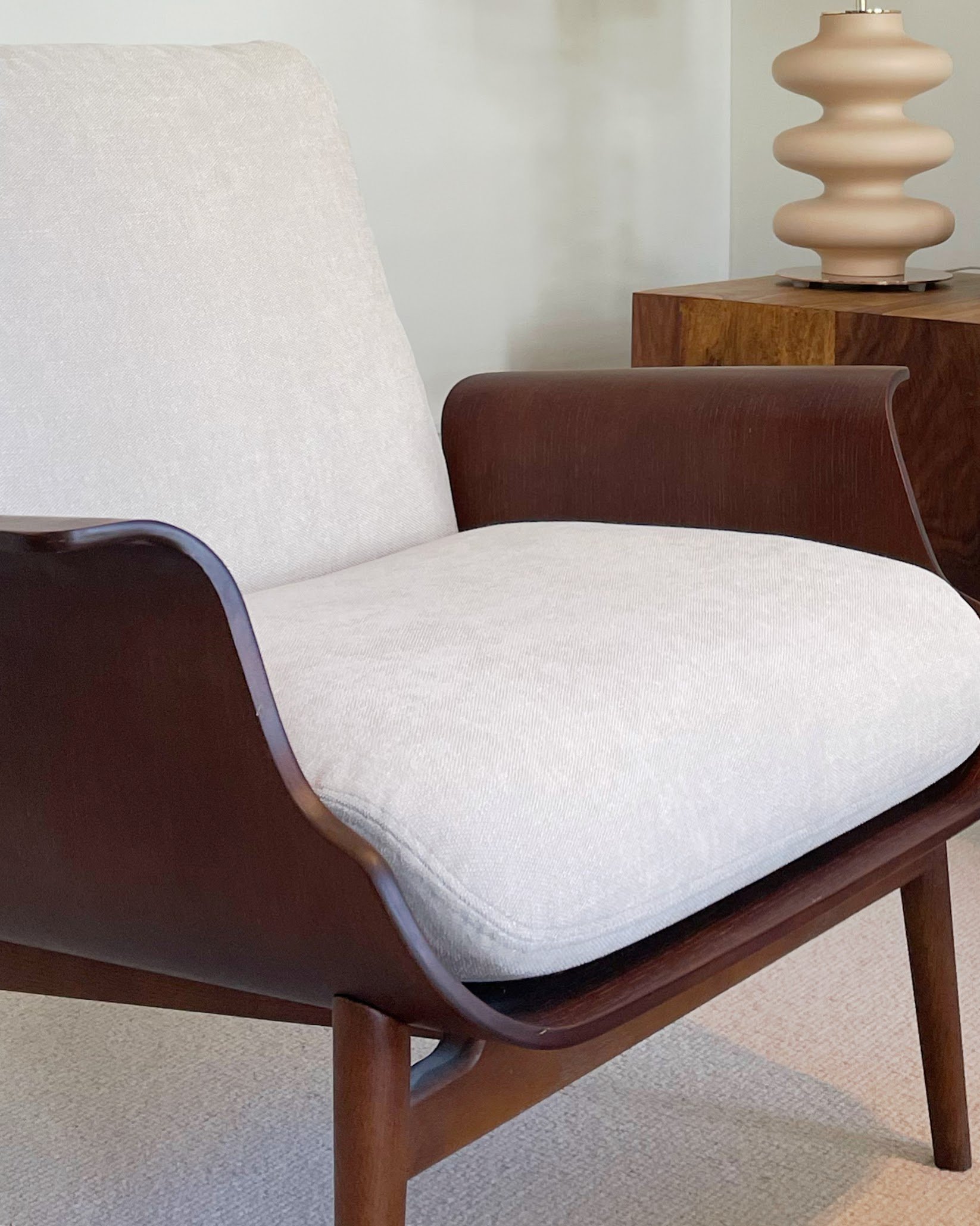 Close-up of a modern mid-century style armchair with cream fabric upholstery and dark wood curved armrests and legs, next to a wooden side table with a textured beige and pink ceramic lamp.