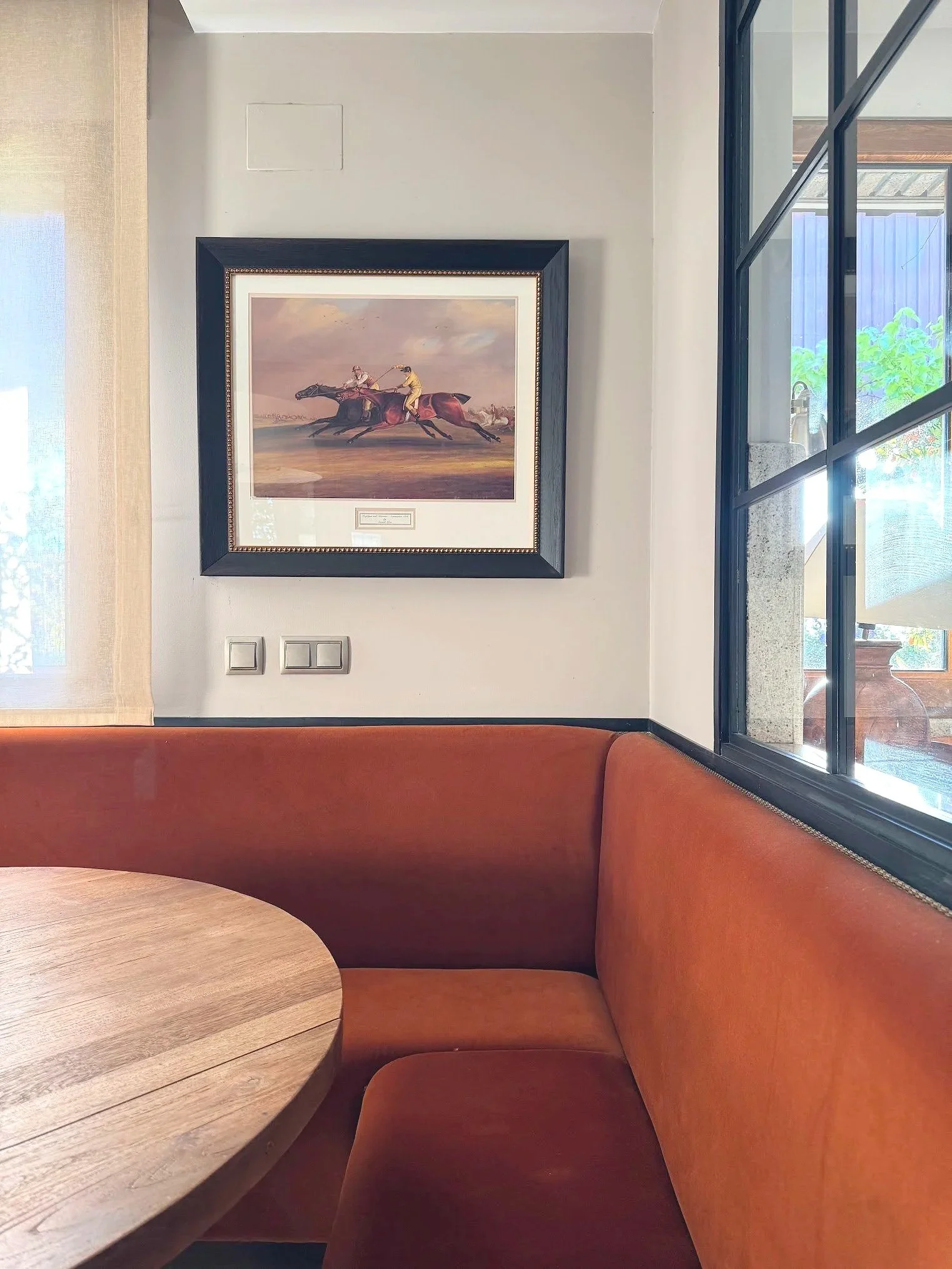 A framed painting of two jockeys racing on horses hangs on a wall above an orange booth seating area, with a wooden table in the foreground and window with a black frame to the right.
