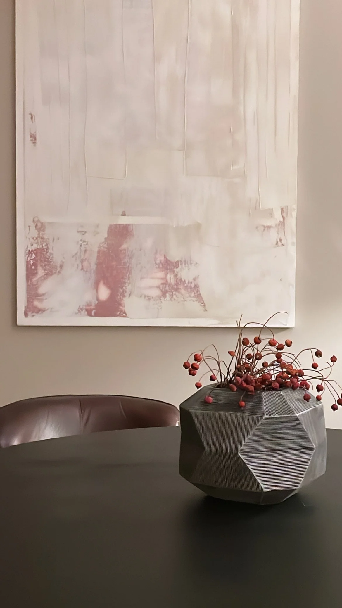 A dark-colored table with a geometric-patterned gray vase containing red berries, with a beige wall, a white textured abstract painting, and a brown leather chair in the background.