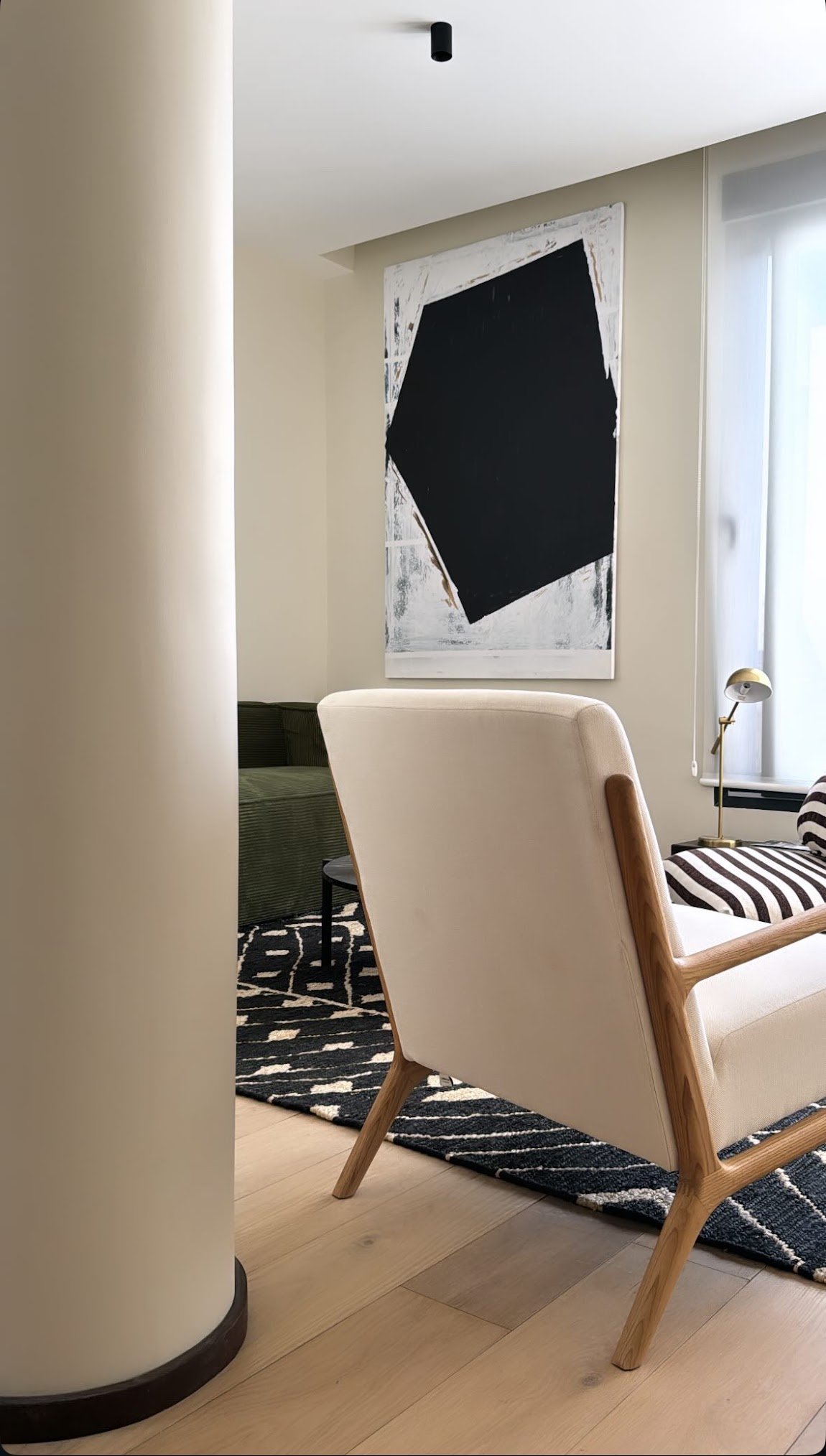 A modern living room corner featuring a white armchair with wooden legs, a black and white patterned rug, a black sofa, a small dark side table, a striped cushion, a brass desk lamp, a large abstract black and white painting, and a sunlight-filled window.