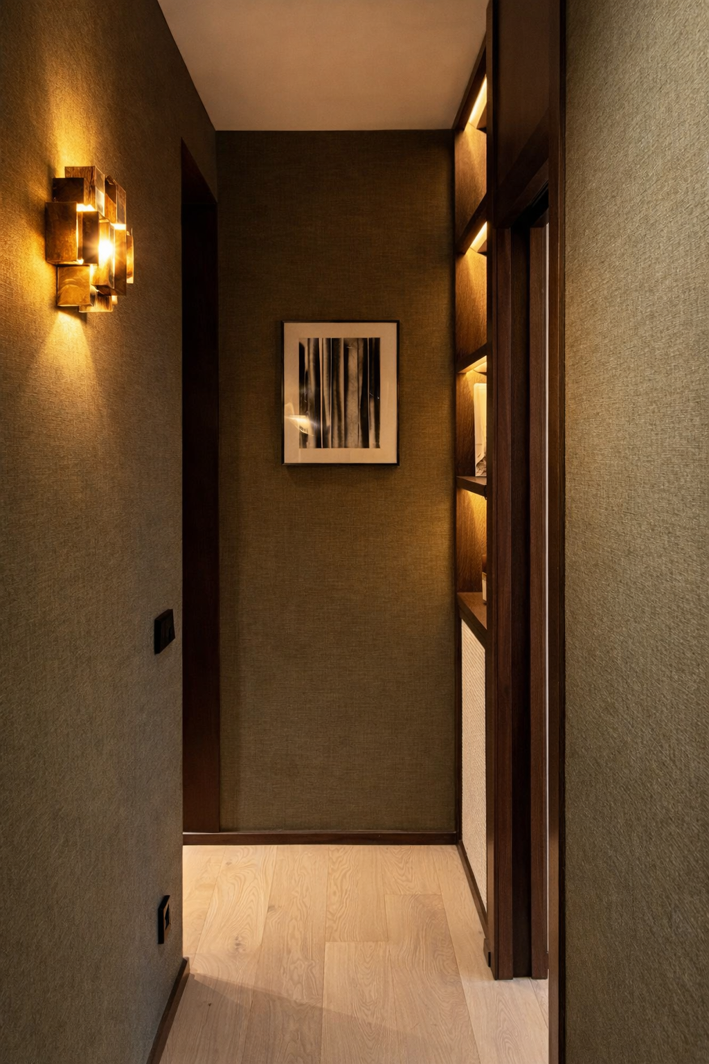 A narrow hallway with textured green walls, a framed black-and-white abstract art piece, a built-in lit bookshelf on the right, and a modern wall sconce with gold tones on the left wall, illuminated with warm lighting.