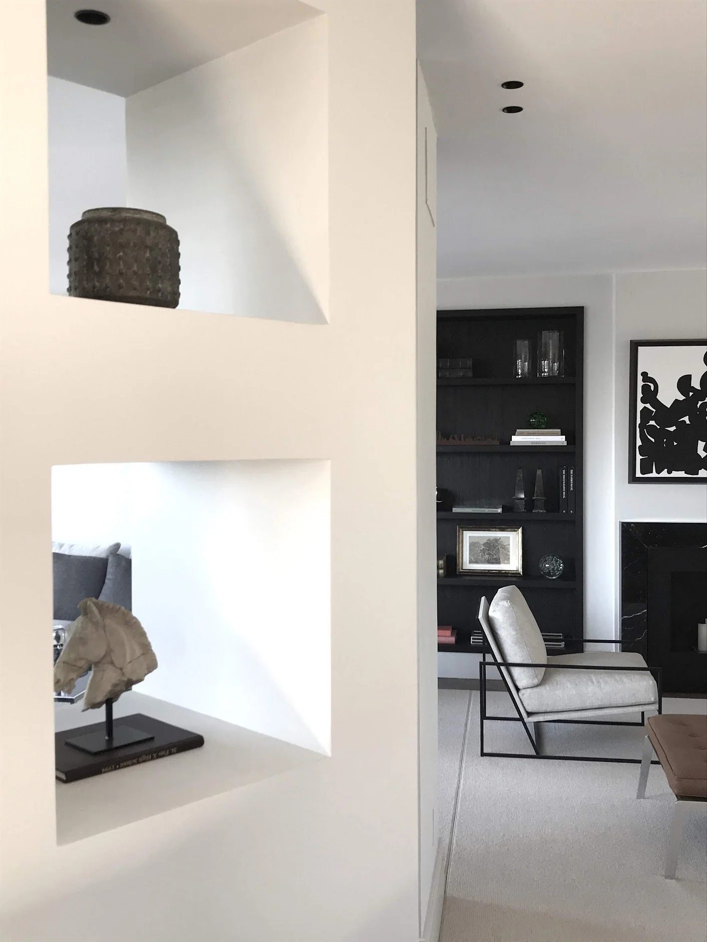 Modern living room with a white wall featuring two square cutouts displaying decorative objects. In the background, there is a black bookshelf with glassware, books, and decorative items, a beige armchair with black metal frame, and a black and white abstract artwork on the wall.