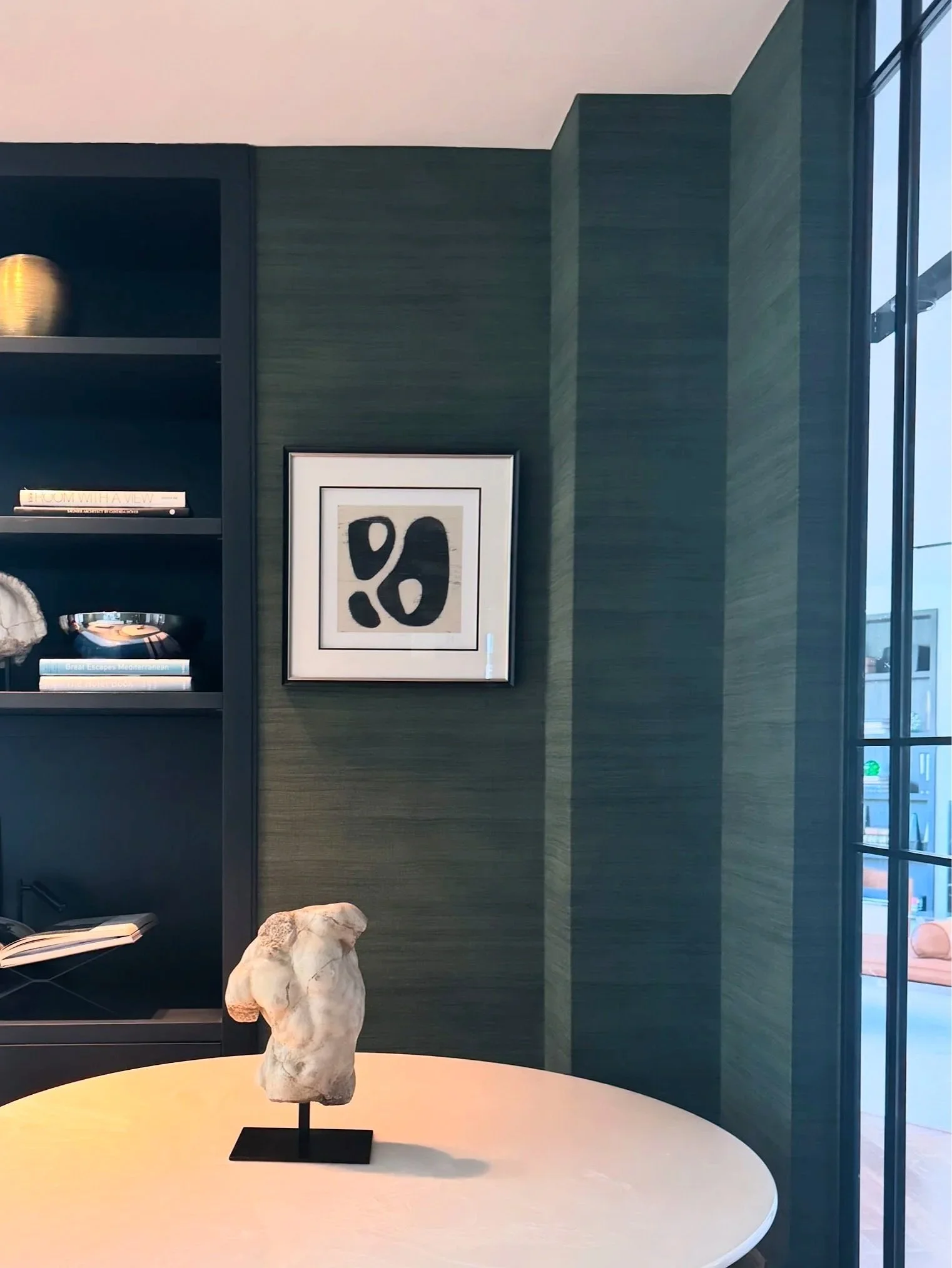 Interior with a dark bookshelf, framed abstract art on green wall, white tabletop with sculpture, and guest room visible through glass wall.