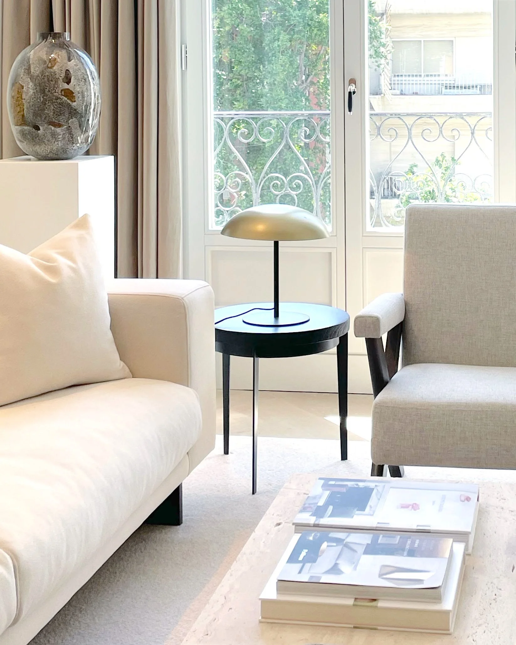 Living room with white sofa, gray armchair, black side table with gold lamp, and glass door leading to balcony with ornate railing, view of trees and neighboring building.