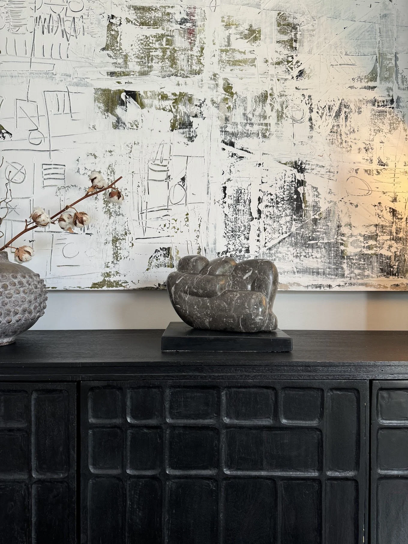 A dark wooden sideboard with a black marble sculpture on top, a gray textured vase with cotton branches, and an abstract white and black art piece on the wall behind.