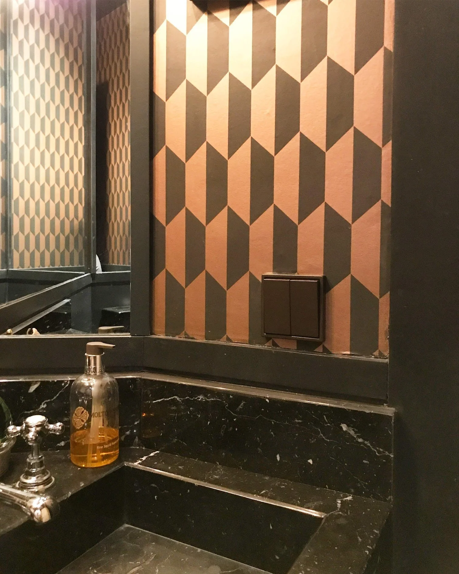 Bathroom with black marble counter, soap dispenser, mirror, geometric patterned wallpaper, and dark-colored switch on the wall.