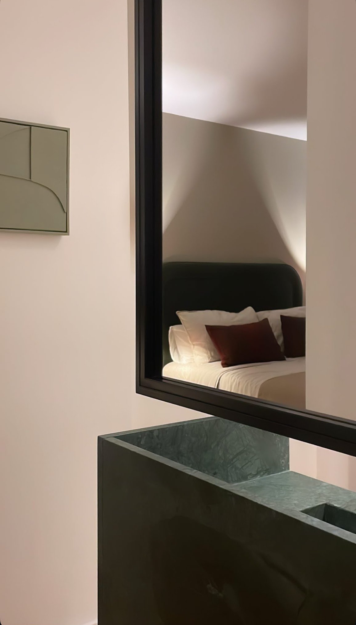 A bedroom reflected in a large mirror, with a bed having white linens and brown pillows, and a sloped ceiling.