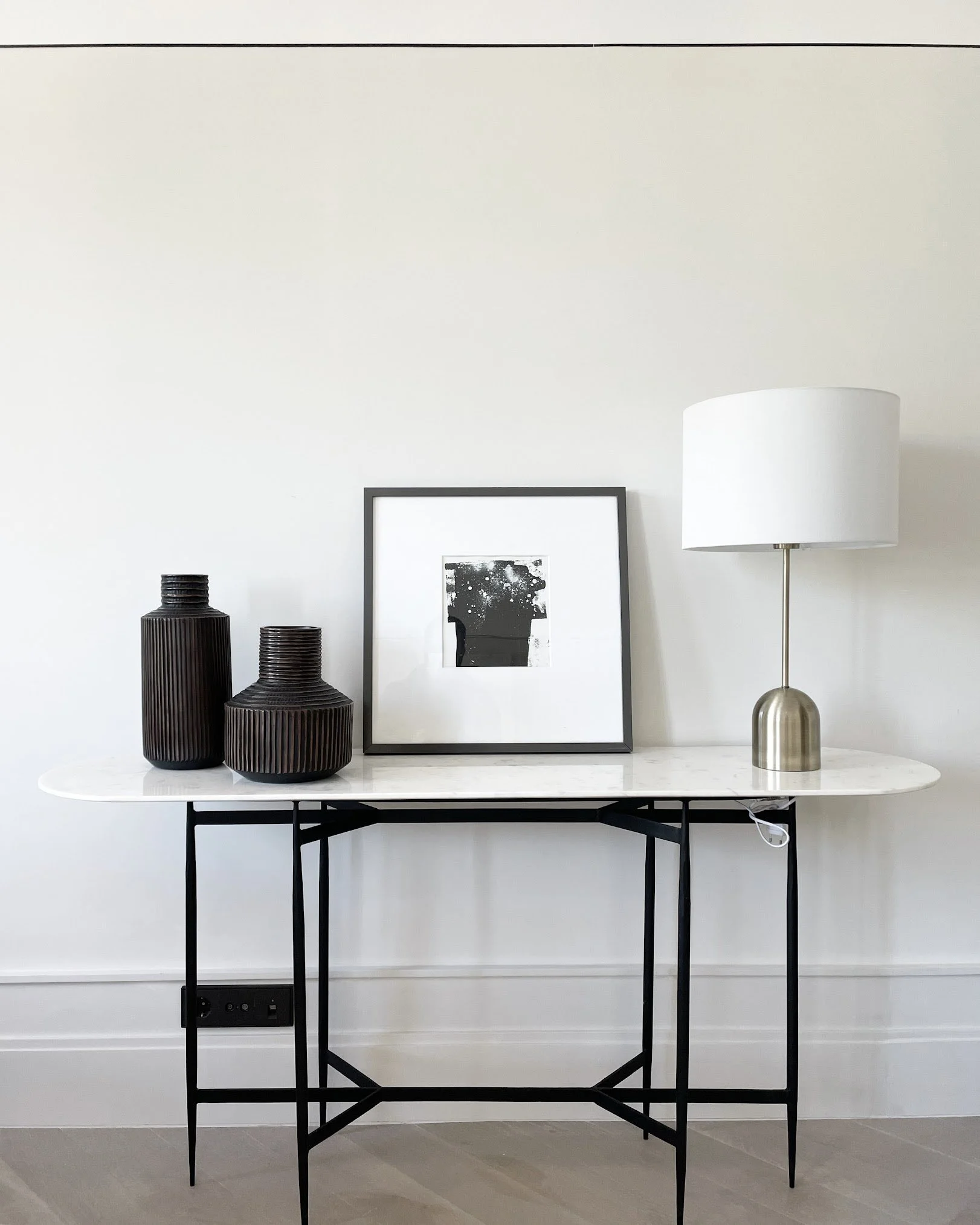 Minimalist console table with black vases, framed black-and-white art, and white lamp against a white wall.