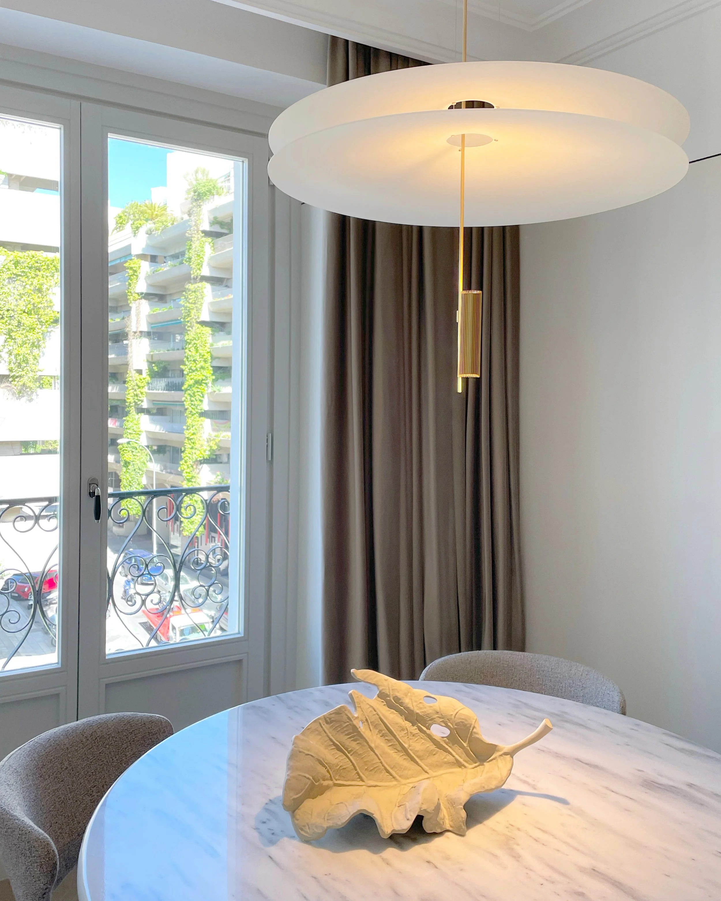 Interior of a modern living room with a round marble table, beige chairs, large window with curtains, a modern hanging light, and a decorative leaf-shaped sculpture on the table.