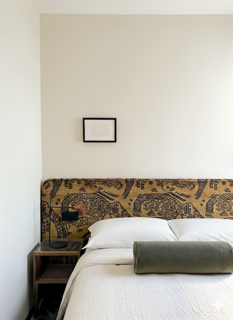 A neatly made bed with white sheets, two pillows, and a green bolster pillow, set against a decorative headboard with African animal print. A small wooden nightstand with a modern reading lamp and a charger is beside the bed, with a framed picture hanging on the plain light-colored wall above.