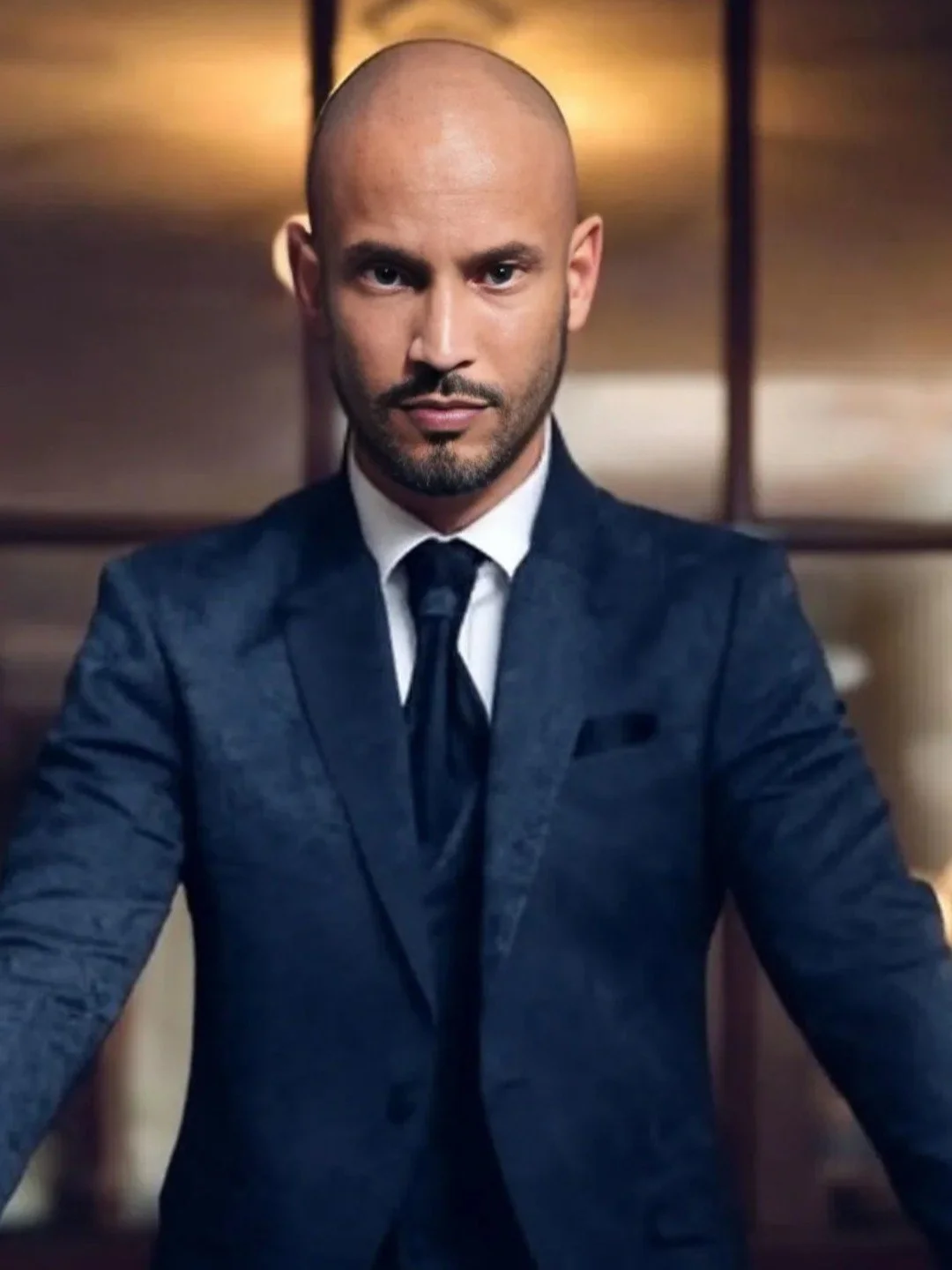 A man in a dark suit and tie with a plaid pattern, no shirt visible underneath, looking directly at the camera in an indoor setting with warm lighting.