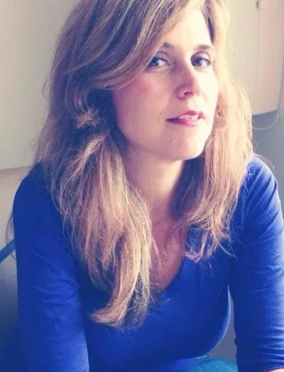 A woman with long, wavy light brown hair wearing a blue top, sitting indoors against a neutral background.