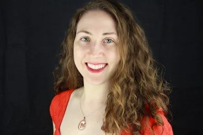 Portrait of a young woman with curly hair, wearing a red top and necklace, smiling against a black background.