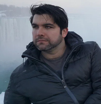 Man with dark hair and beard wearing a black jacket outdoors near a body of water