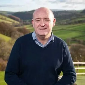 Waist shot photo of John Davies, owner of Beacons View self catering lodges and cottages in the Brecon Beacons wales, with lush, green farmland in the background.jpg