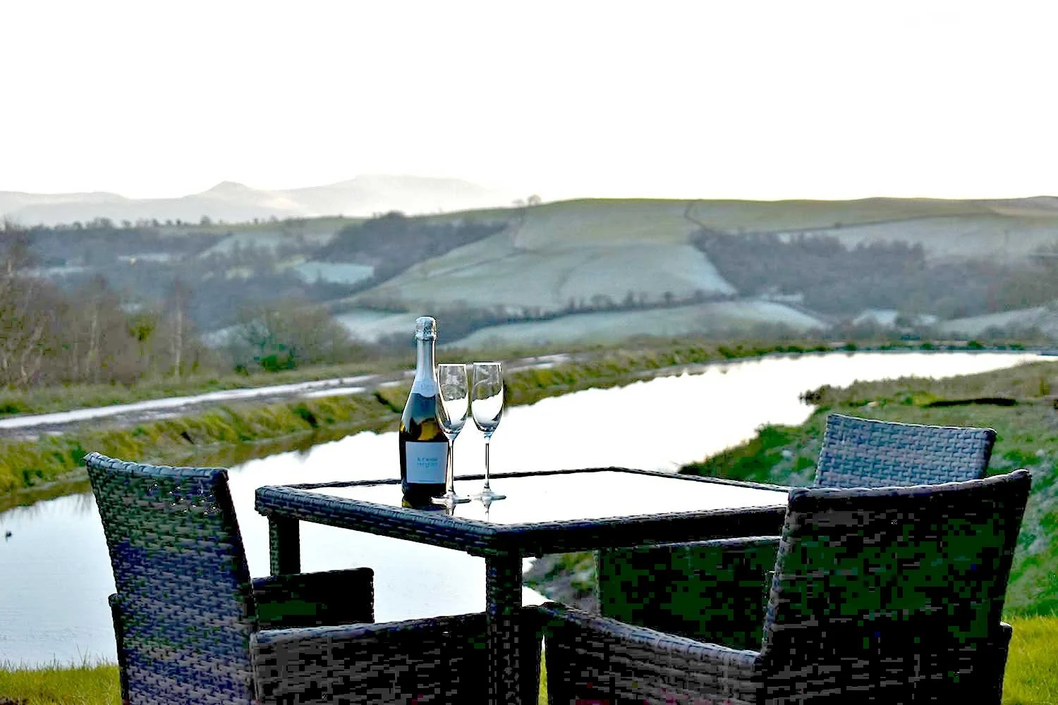 A table with two champagne glasses and a bottle set outdoors overlooking a lake and Brecon Beacons in the distance.