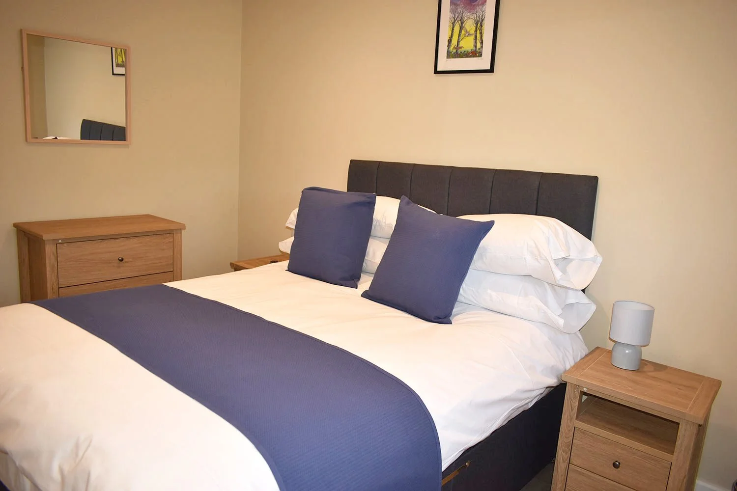 A neatly made bed with white sheets, a blue bed runner, and two blue pillows in a bedroom with wooden furniture and a framed picture of trees on the wall.