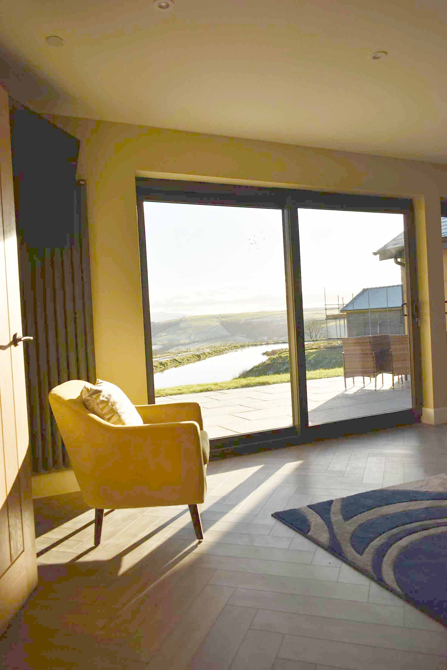 Living room with a yellow armchair, patterned rug, and a large glass sliding door leading to an outdoor patio with a view of a river, grassy area, and rolling hills in the distance.