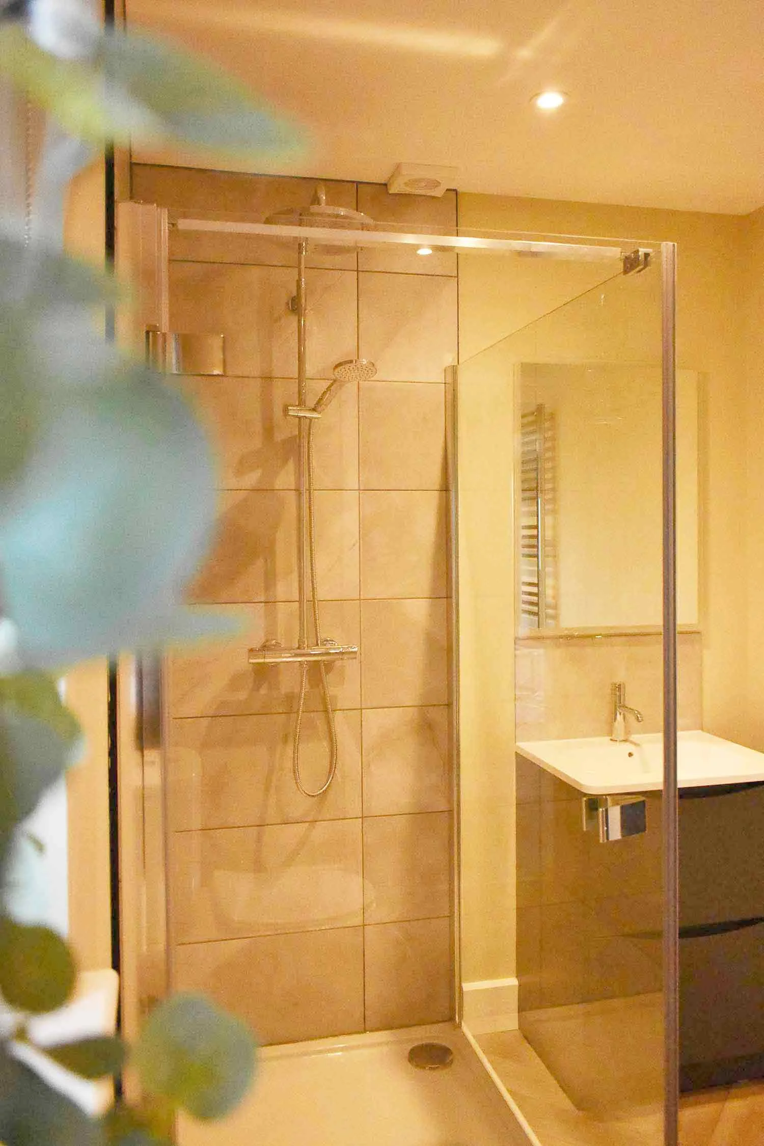 A modern bathroom at Beacons View lodges in the Brecon Beacons, with a walk-in shower, a white sink, and a mirror. The shower has beige tiles and a glass partition. There are recessed ceiling lights.
