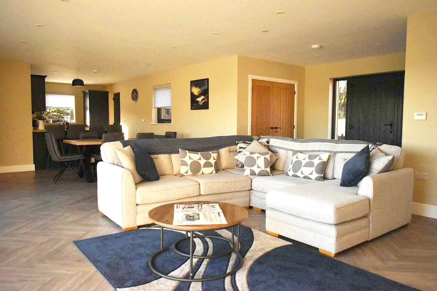 Beacons View lodge living room with beige sectional sofa, patterned pillows, wooden coffee table on a blue area rug, open floor plan with dining area and kitchen, wooden door, black sliding barn door, and small windows with curtains.