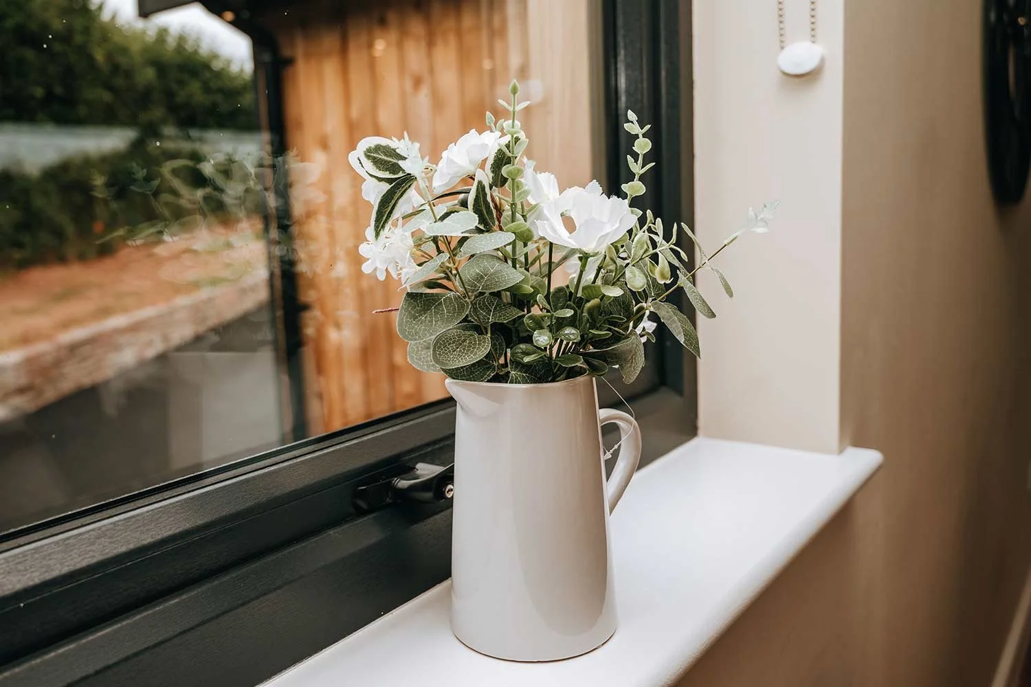 A white ceramic vase with white flowers and green leafy foliage on a windowsill.