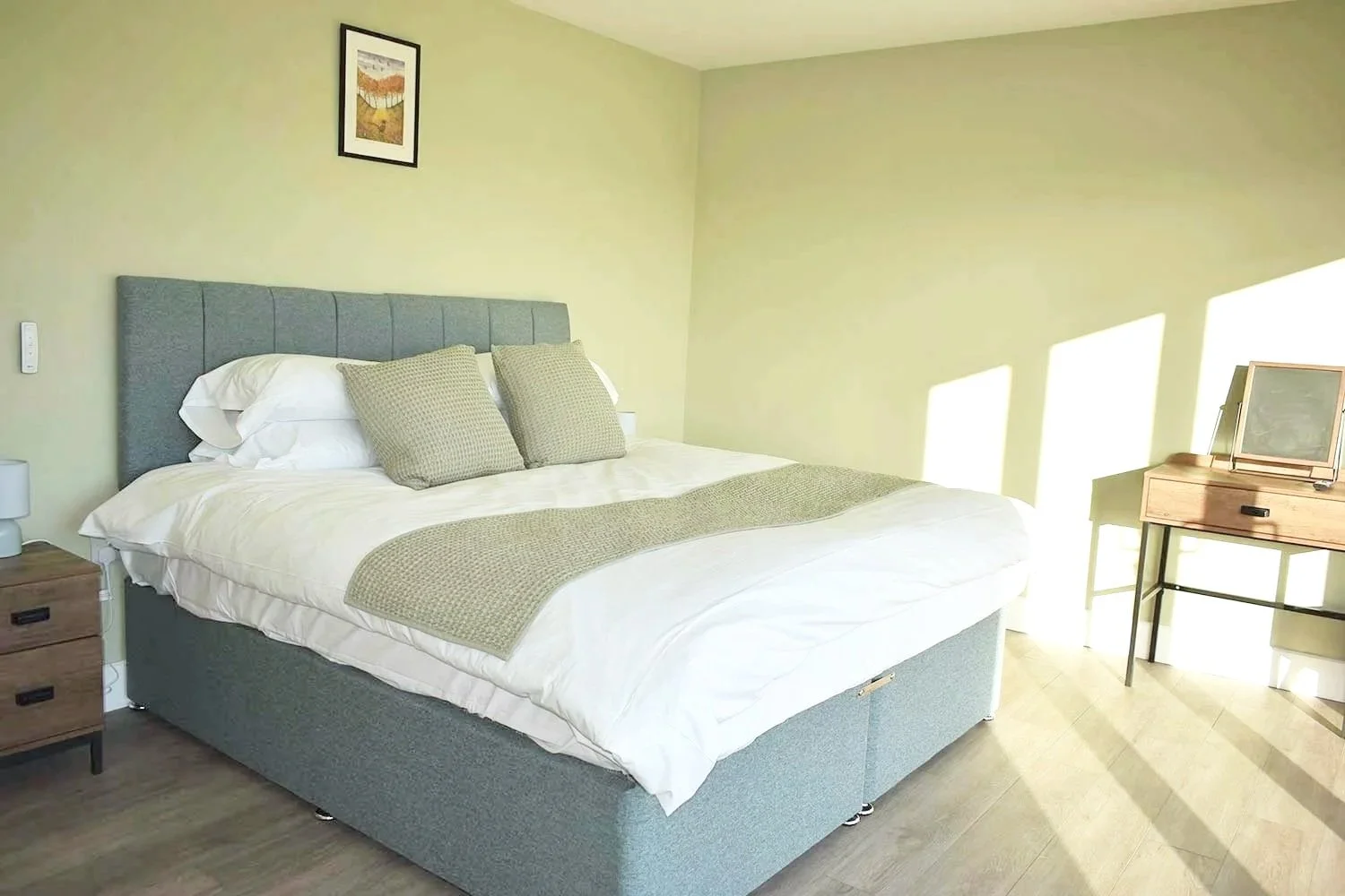 A bedroom with a large bed featuring a gray headboard, white bedding, and beige pillows. There are two wooden nightstands and a wooden desk near a window with bright sunlight streaming in.