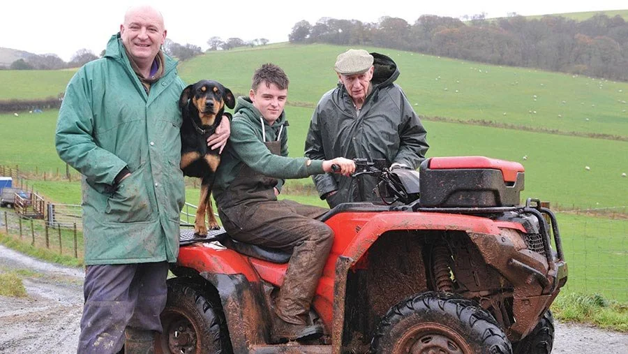 John and Elwyn Davies stood either side of Brychan Davies on a red quad bike with a dog on the back, taken outside with stunning Brecon Beacons farmland in the background.