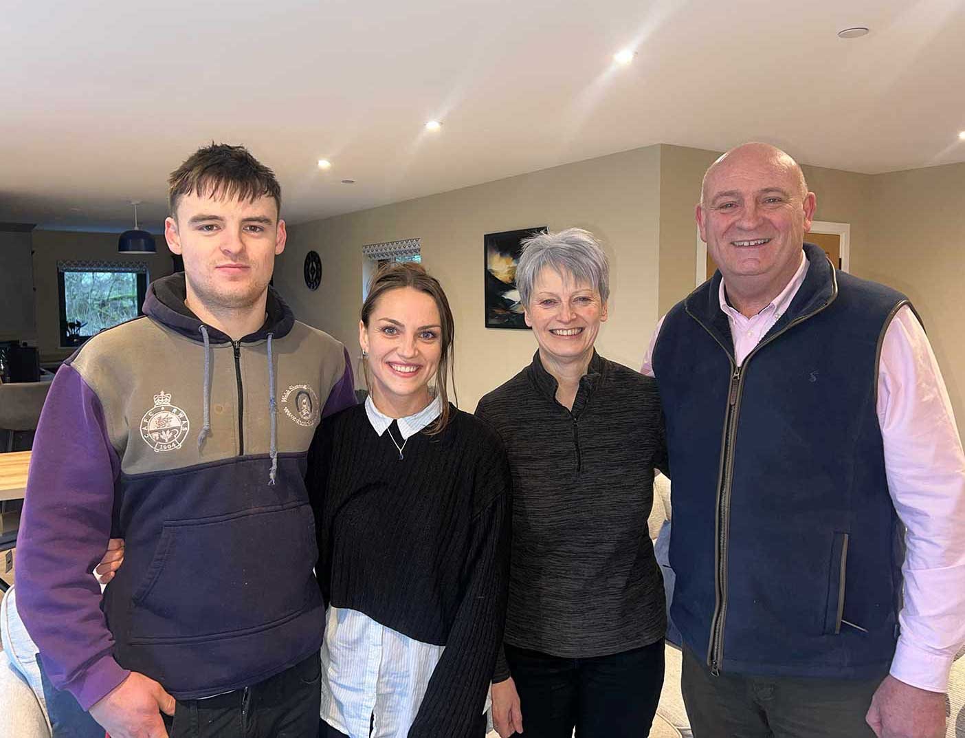 A smiling group of four, Brychan, Shauna, Menna and John, standing together in the modern, open-plan interior of Beacons View luxury holiday lodge in the Brecon Beacons.