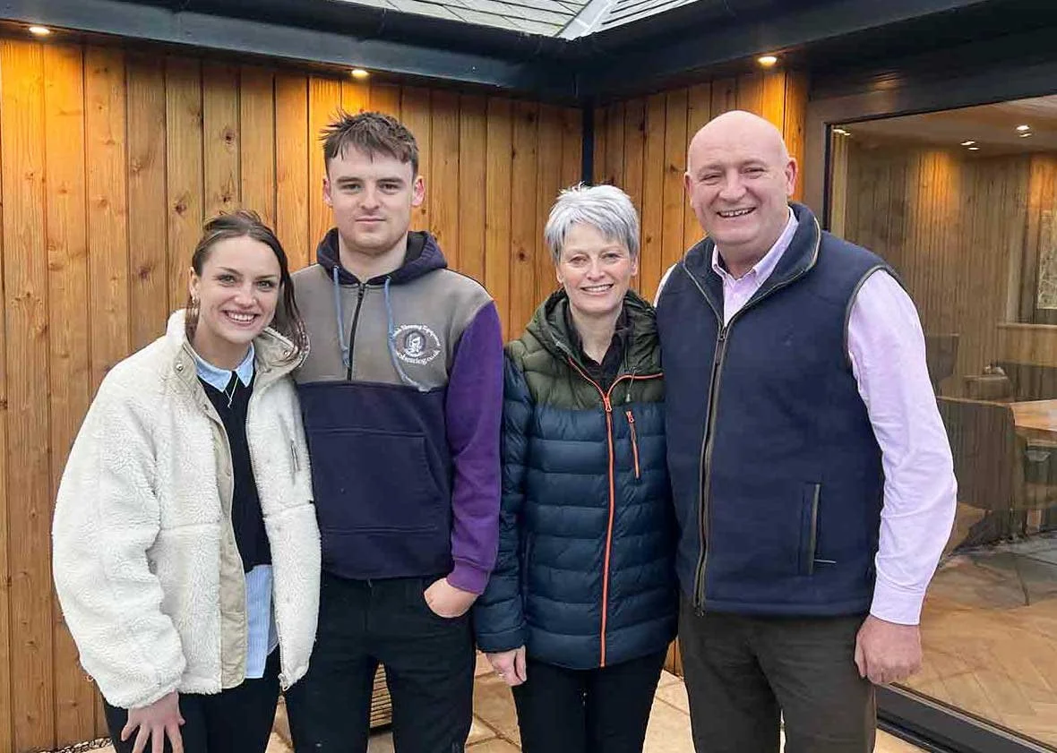 Shauna, Brychan, Menna and John standing outdoors in front of the wooden wall of one of their luxury self-catering lodges in the Brecon Beacons in Wales, smiling at the camera and dressed in casual outerwear.