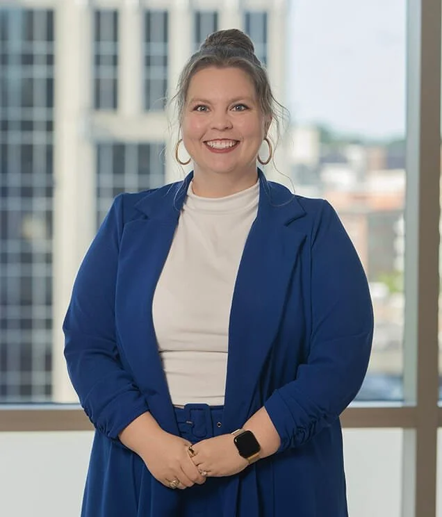 A smiling woman wearing a blue blazer, white shirt, and a smartwatch, standing in front of large windows with city buildings in the background.