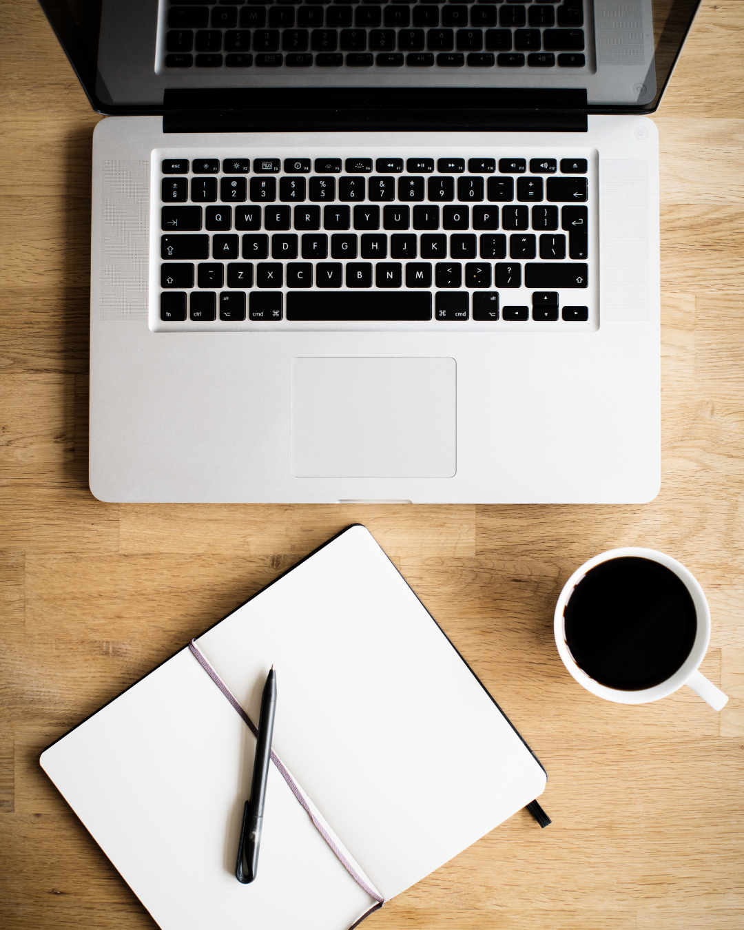 Top-down view of a workspace with a laptop, an open notebook with a pen on it, and a cup of black coffee on a wooden table.