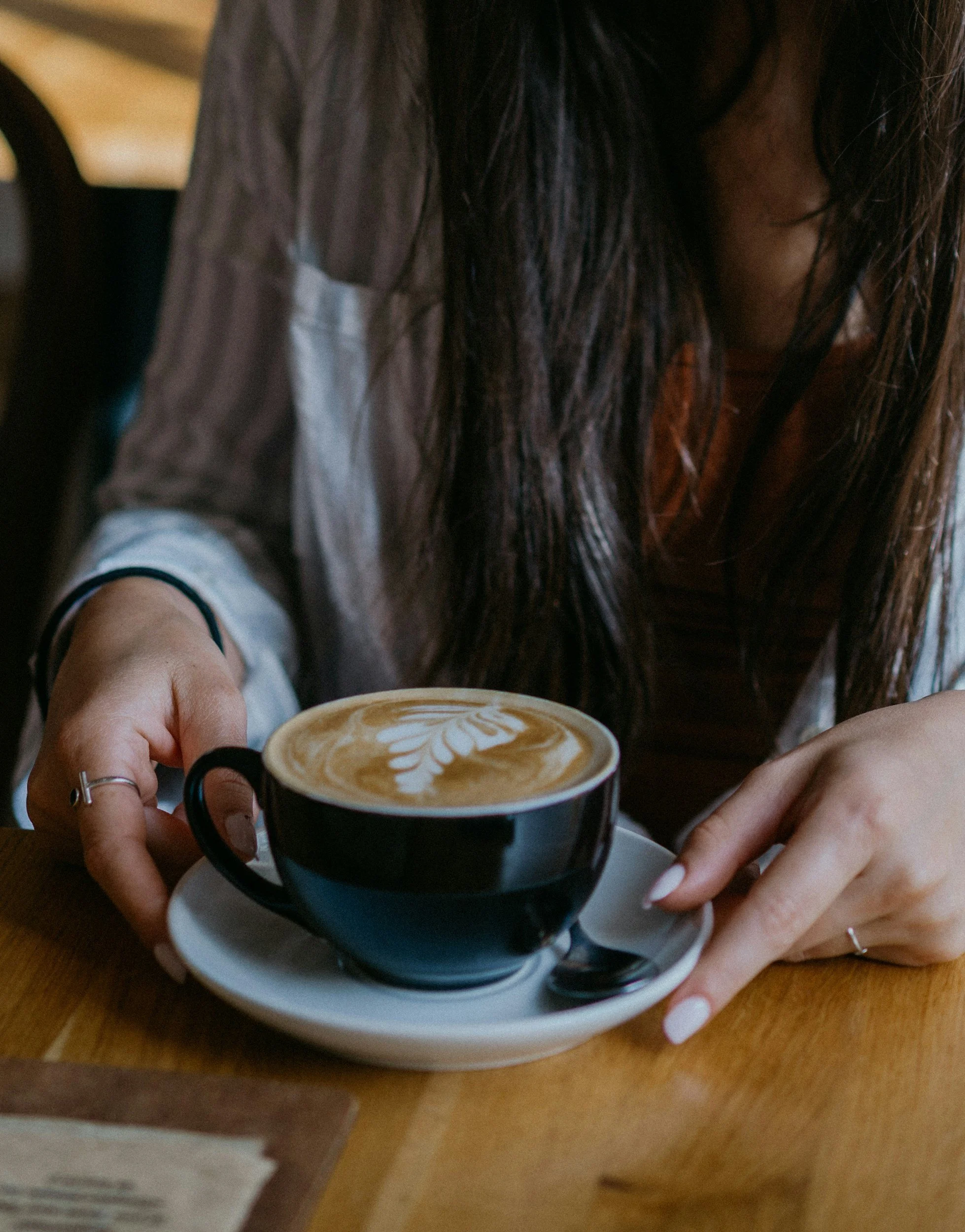 Person with long dark hair holding a black coffee cup with latte art, sitting at a wooden table in a cafe.