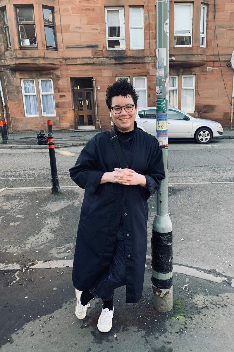 A person with short curly hair, glasses, and a dark coat standing on a city street next to a lamppost with stickers. They are smiling and looking at the camera.