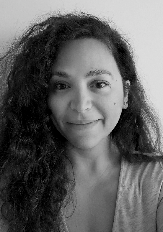 A black-and-white photo of a woman with long, curly hair and a slight smile, wearing a light-colored top.
